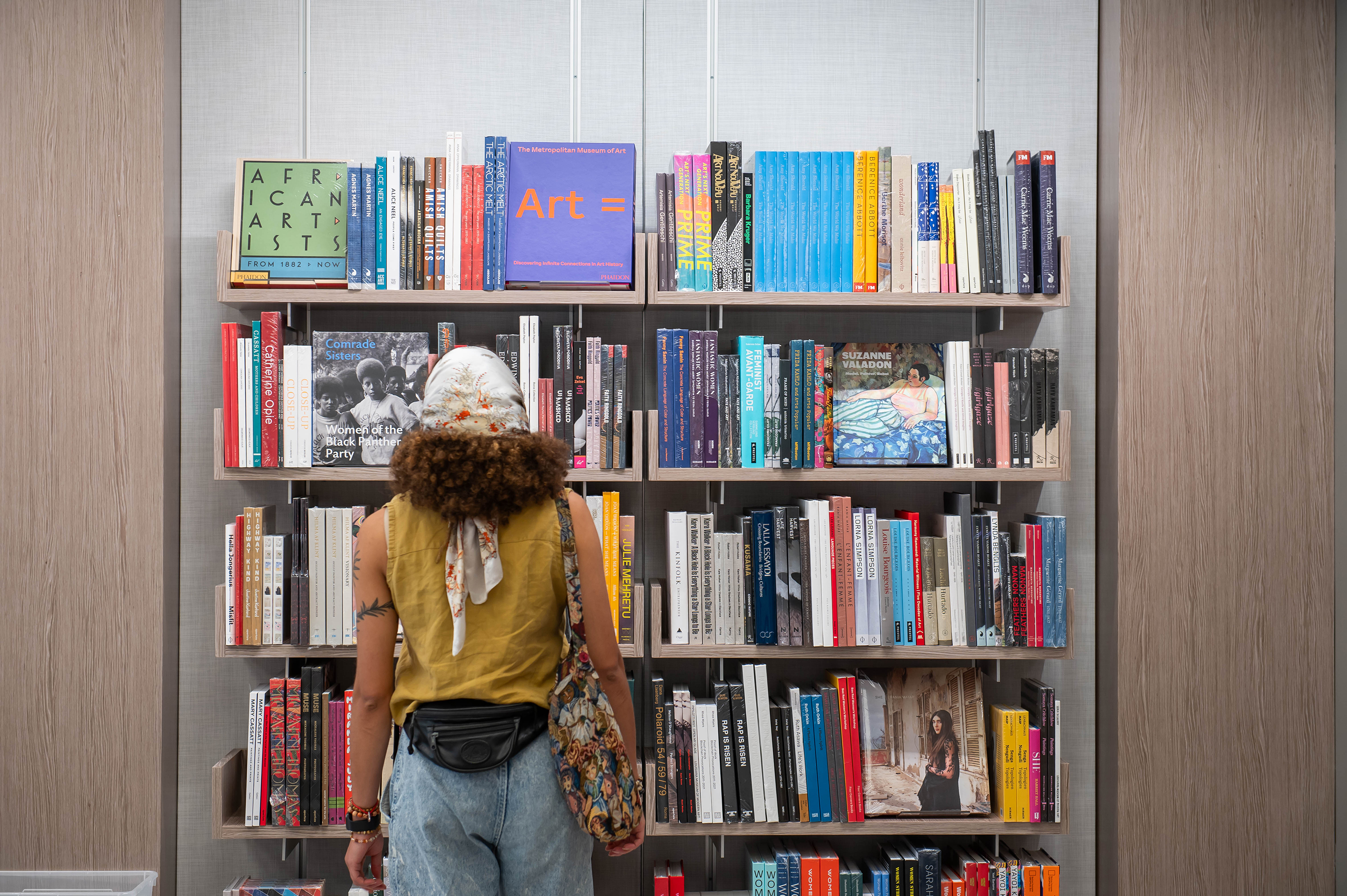 A visitor with medium skin tone and brown curly hair wearing a white and red floral head scarf, ochre sleeveless shirt, black backwards fanny pack, multicolored tote bag, and jeans stands facing a large floating bookcase filled with art books and catalogues.