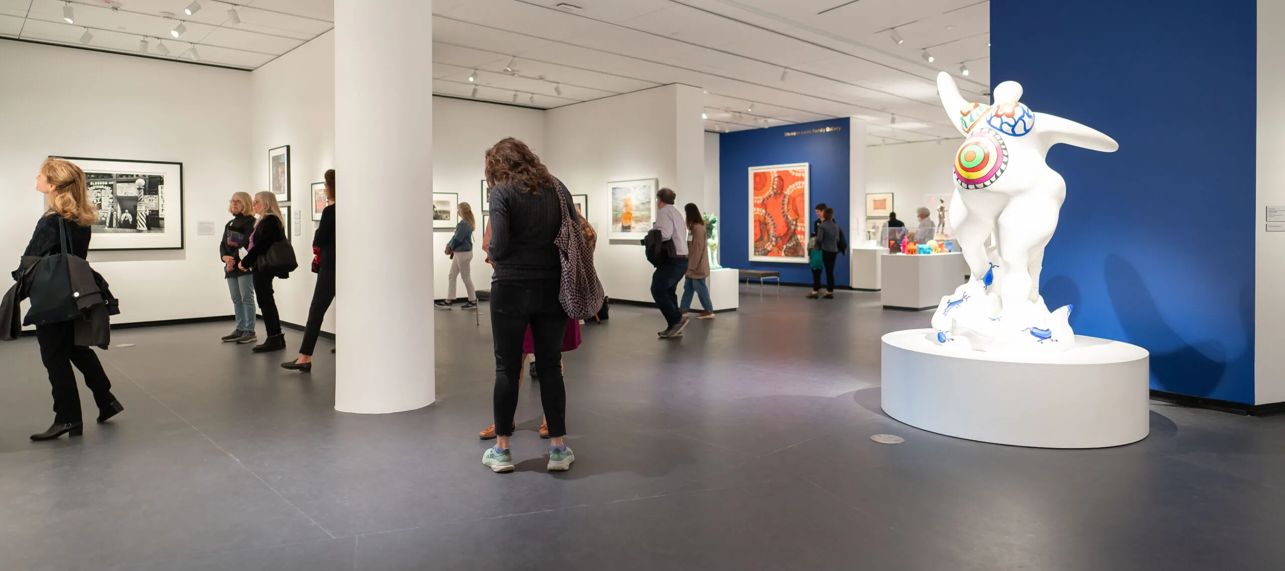 A modern museum gallery is photographed at a wide angle. Visitors observe large artworks on the walls and sculptures on pedestals.