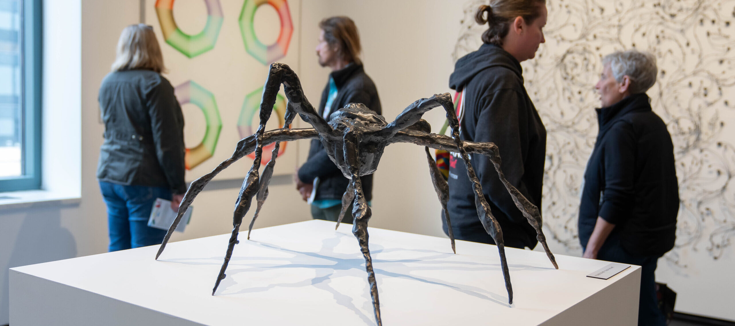 A bronze sculpture of a spider with arched, spindly legs sits on a white pedestal in a museum gallery. In the background, museum visitors observe artworks on the walls.