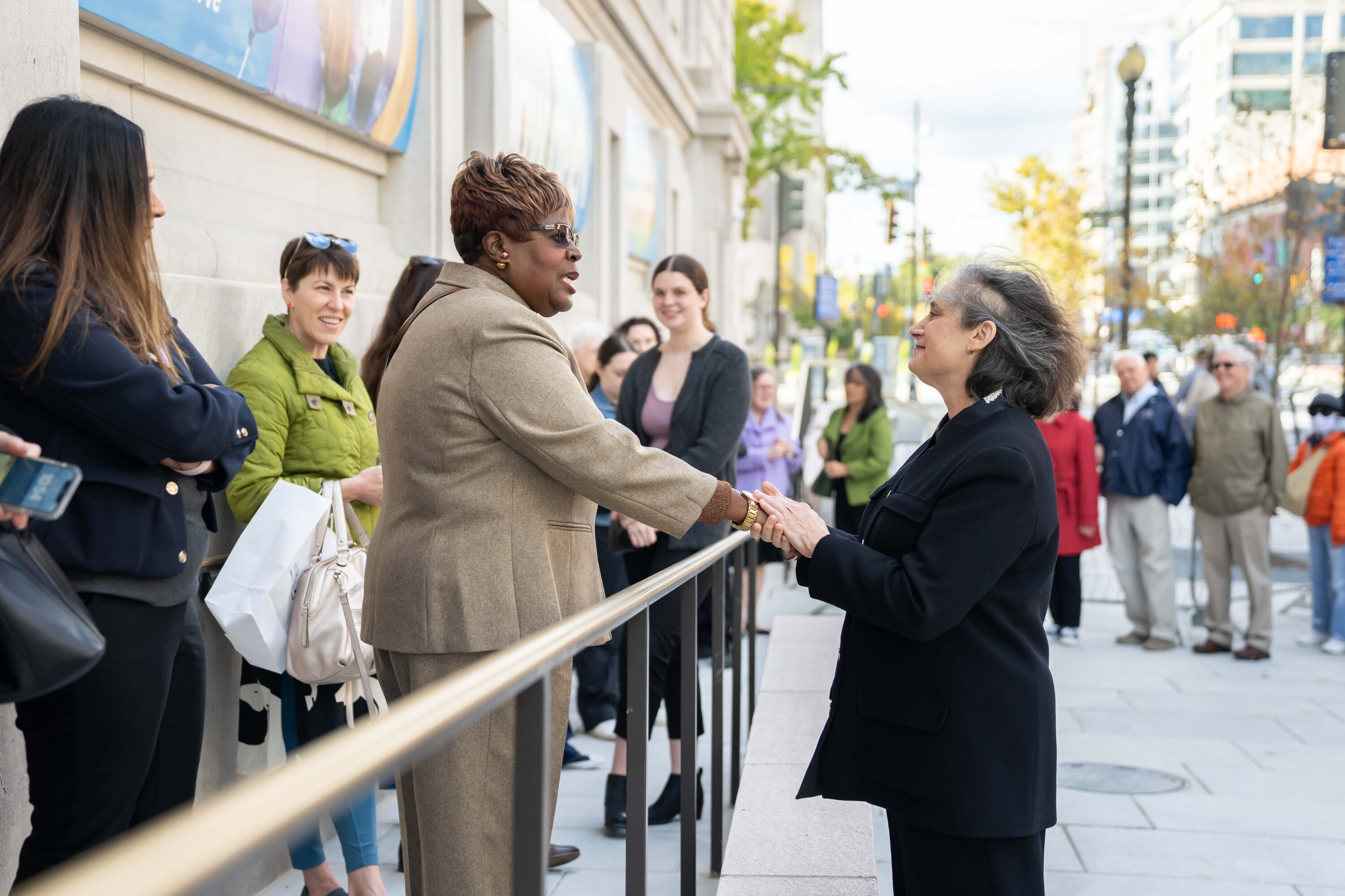 A group of people are standing in line outside of a building. A woman with a light skin tone and gray hair is shaking the hands of a woman standing in line. The woman standing in line has a dark skin tone and is wearing a beige suit.