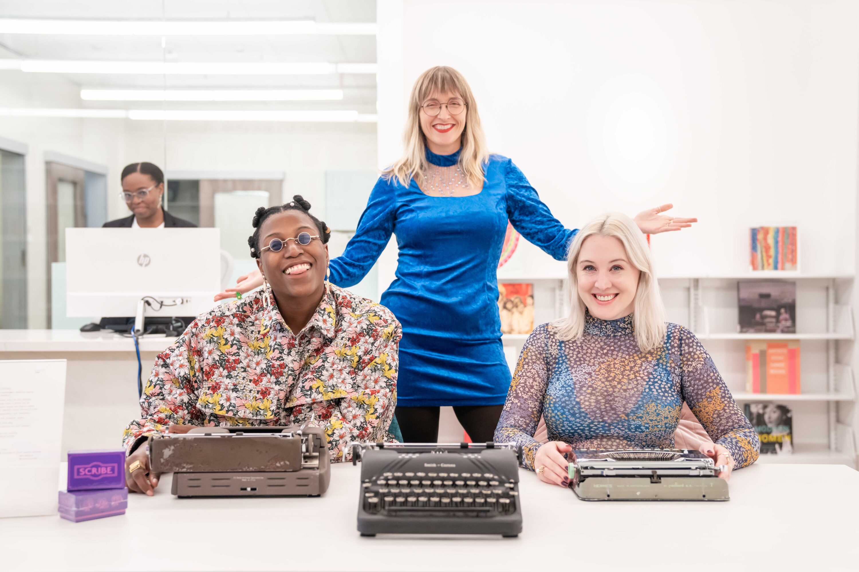 Four women are sitting and standing in a bright and airy room. In front of them are three type writers, which two of the women are using. The women have dark and light skin tones and are wearing floral shirts and velvet dresses. All of them are smiling at the camera. The woman in the back is sitting at a computer screen.