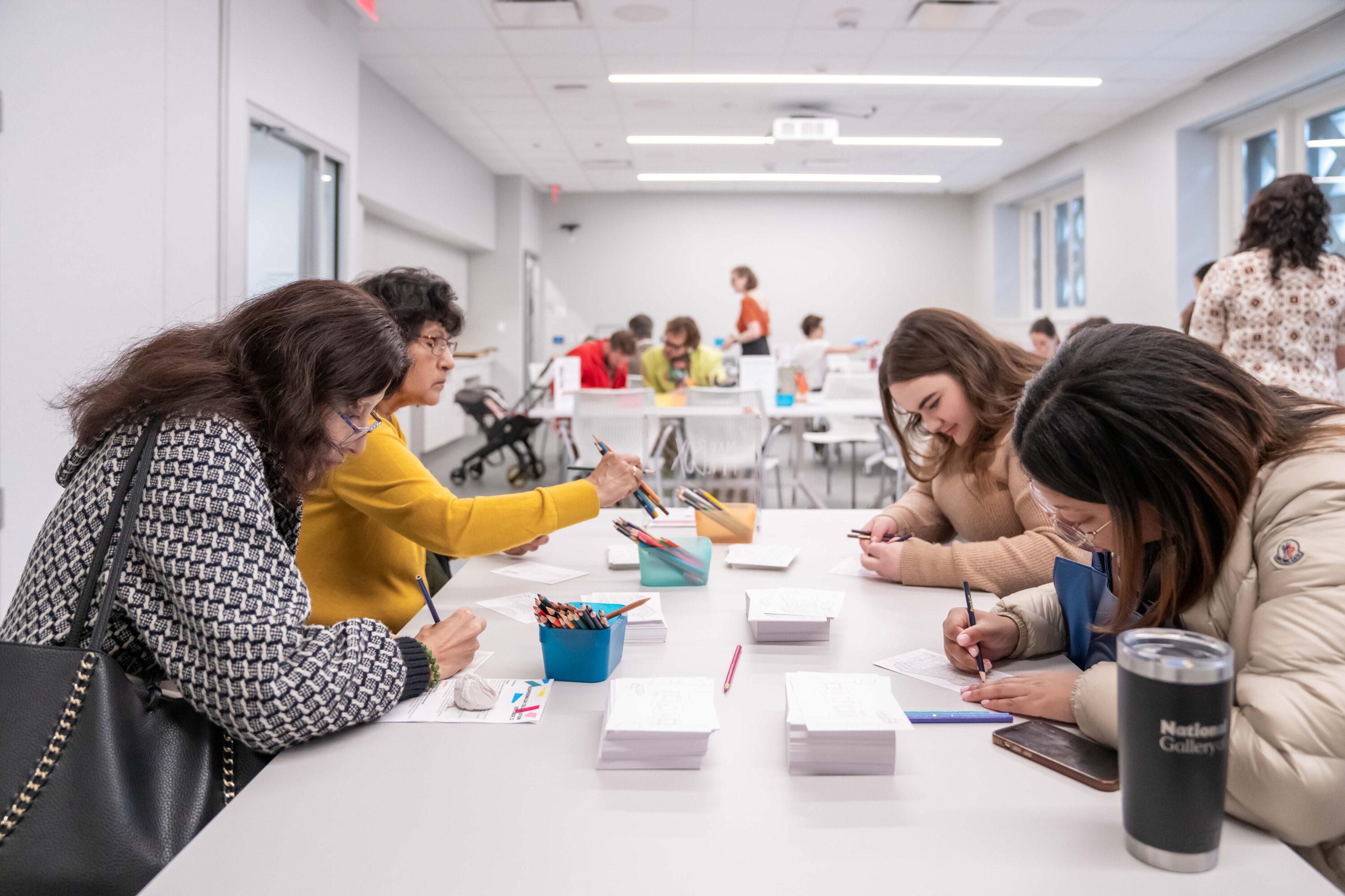 Four women of different ages sit at a white table in a large studio/classroom space. They use colored pencils to color a postcard-size paper. In the background many other people are in the room at other tables doing the same.