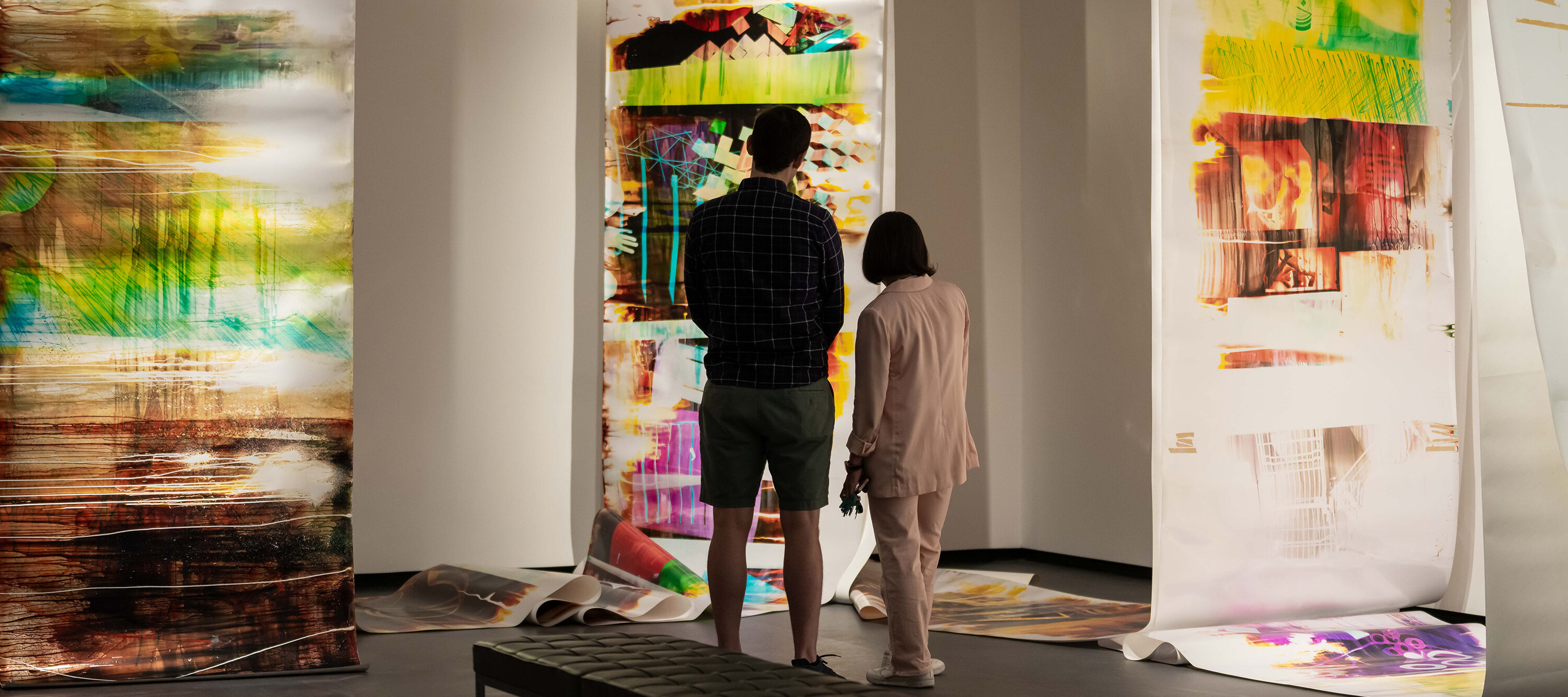 Two museum visitors are standing in front of an artwork. This large-scale, scrolling, colorful, abstract photograph is hung from the ceiling throughout the room.
