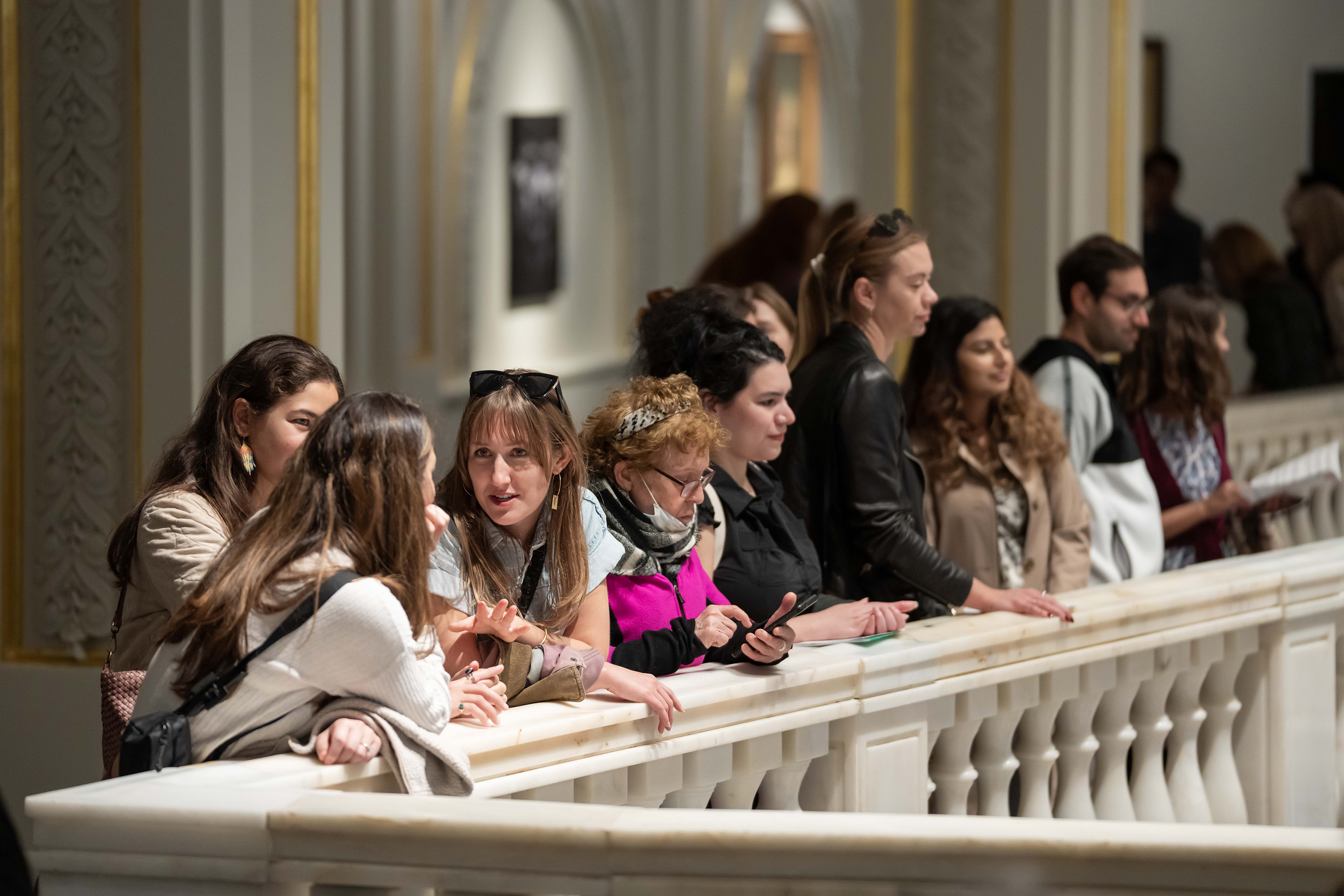 Nine museum visitors of various ages congregate on a marble railing, some chatting, one looking at their phone, and others looking out over the railing.