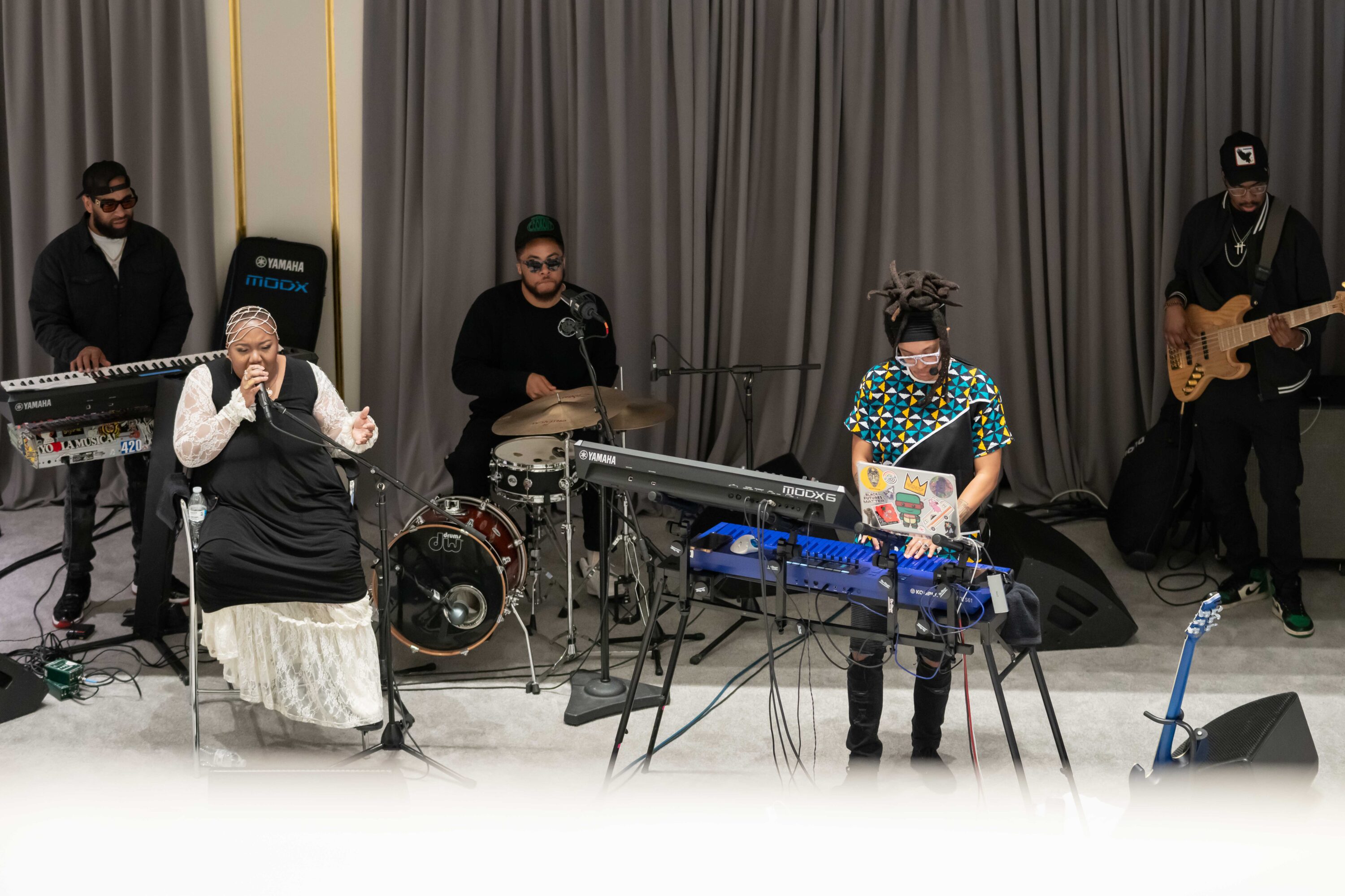 A five-piece band performs on a stage. The three backing members are all men wearing black trendy street wear. The two leading members, positioned in the front, are Black women; one sings and the other plays on a blue keyboard.