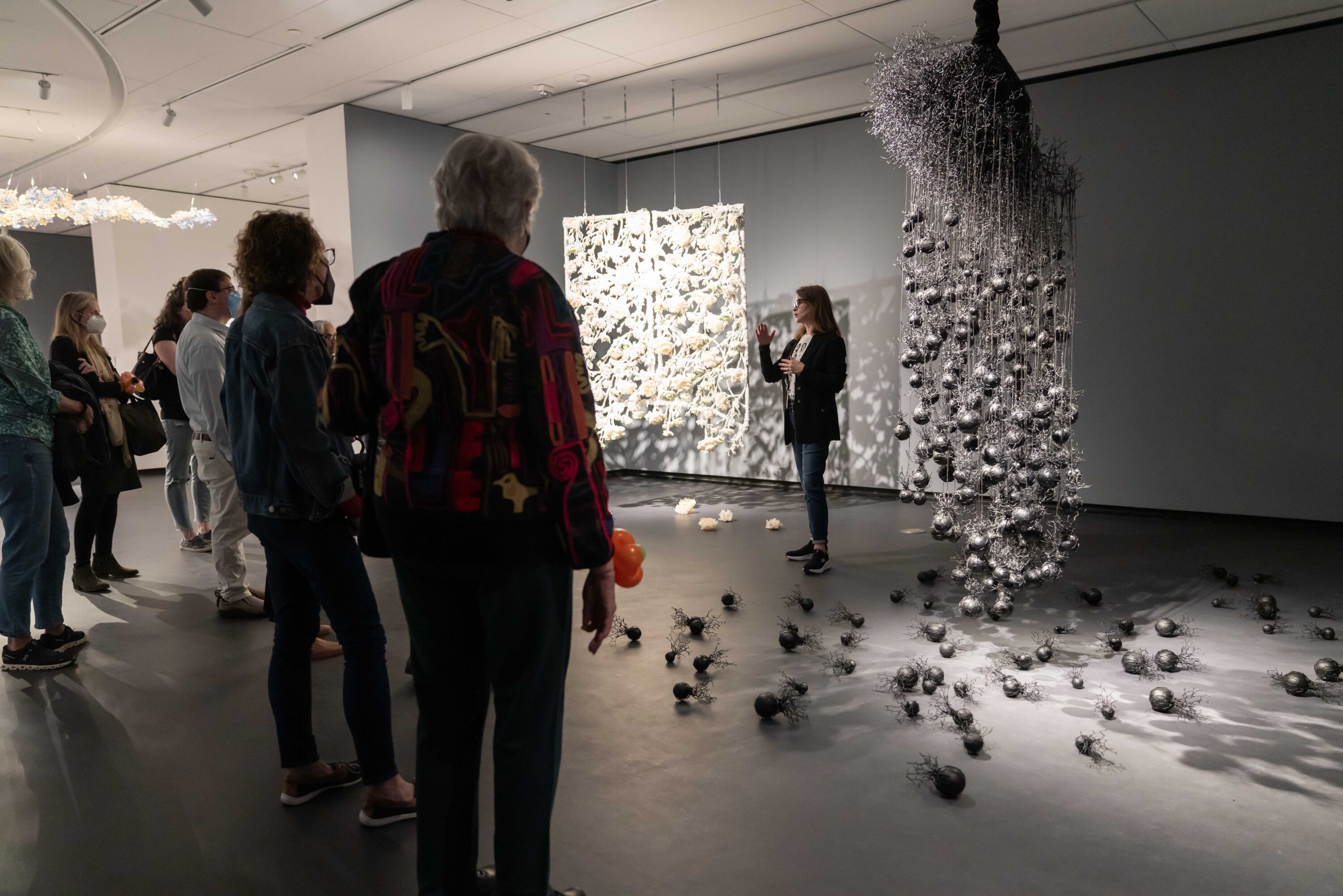 In a dimly-lit museum gallery, a light-skinned woman stands between two large abstract wax sculptures that hang from the ceiling. One is a a white intricate rectangle of connected flowers and vines; the other is a black cluster of fine, vein-like threads and hanging spheres. Below each sculpture, are more of their elements are arranged on the ground. The woman faces a group of visitors who look on.