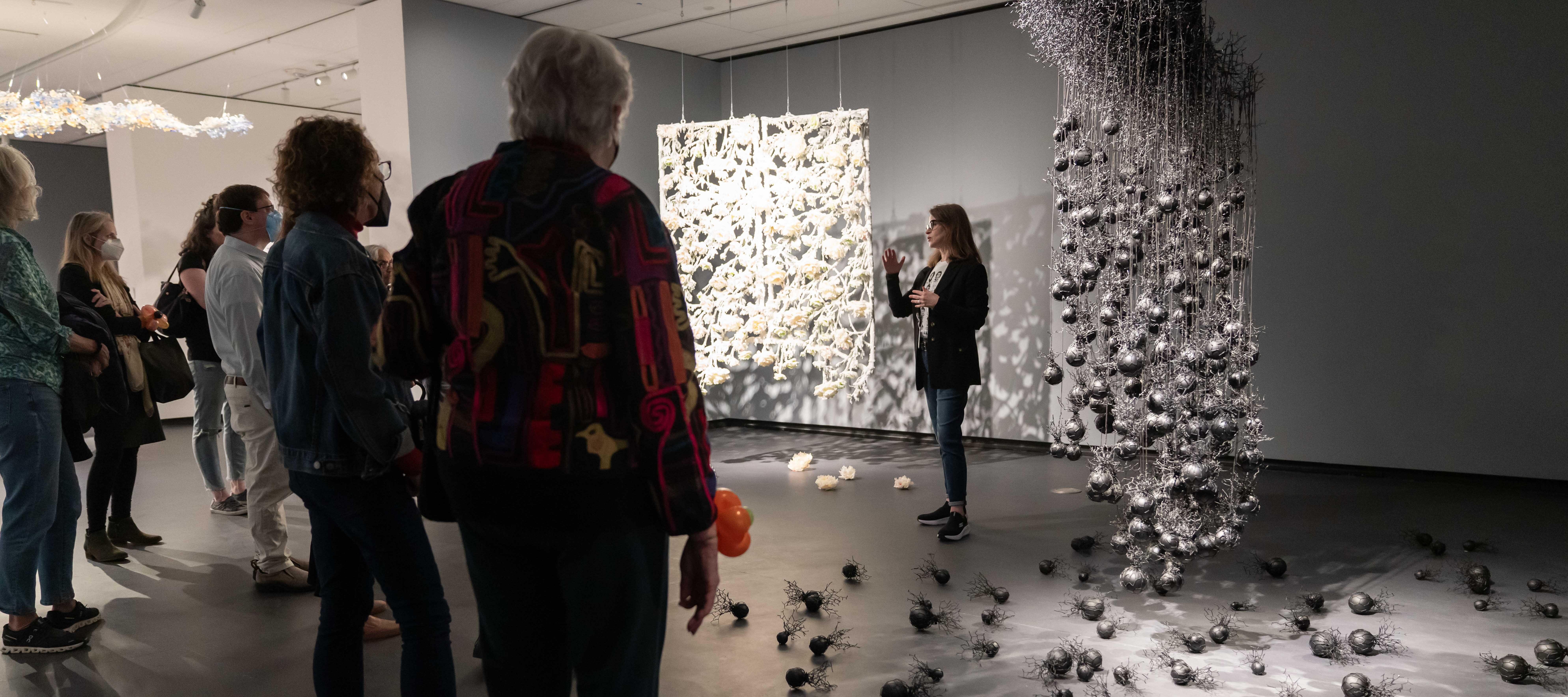 In a dimly-lit museum gallery, a light-skinned woman stands between two large abstract wax sculptures that hang from the ceiling. One is a a white intricate rectangle of connected flowers and vines; the other is a black cluster of fine, vein-like threads and hanging spheres. Below each sculpture, are more of their elements are arranged on the ground. The woman faces a group of visitors who look on.