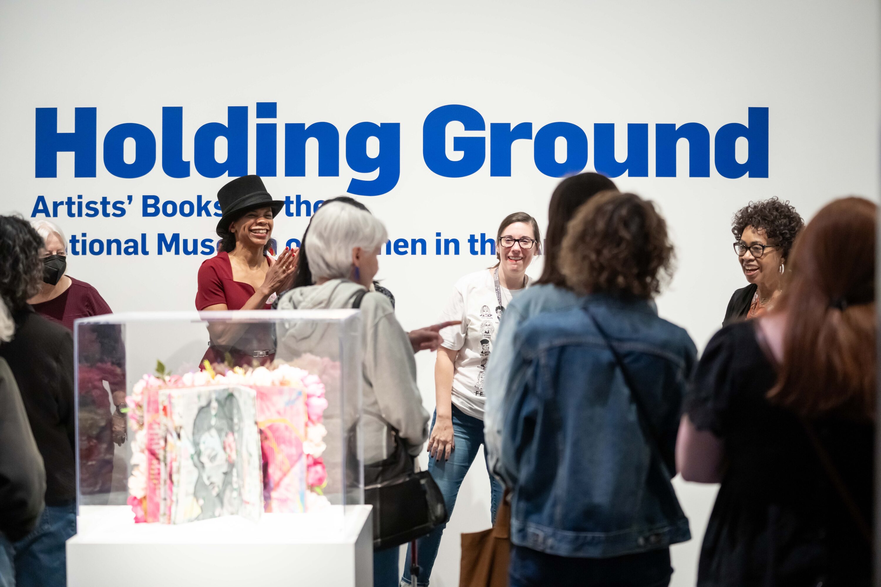 Many people of different ages and races stand in a white-walled museum gallery, some smiling and clapping. A display case containing an artist's book is in the center of the room. Behind everyone, on the wall, is large blue text that reads "Holding Ground: Artists' Books for the National Museum of Women in the Arts."