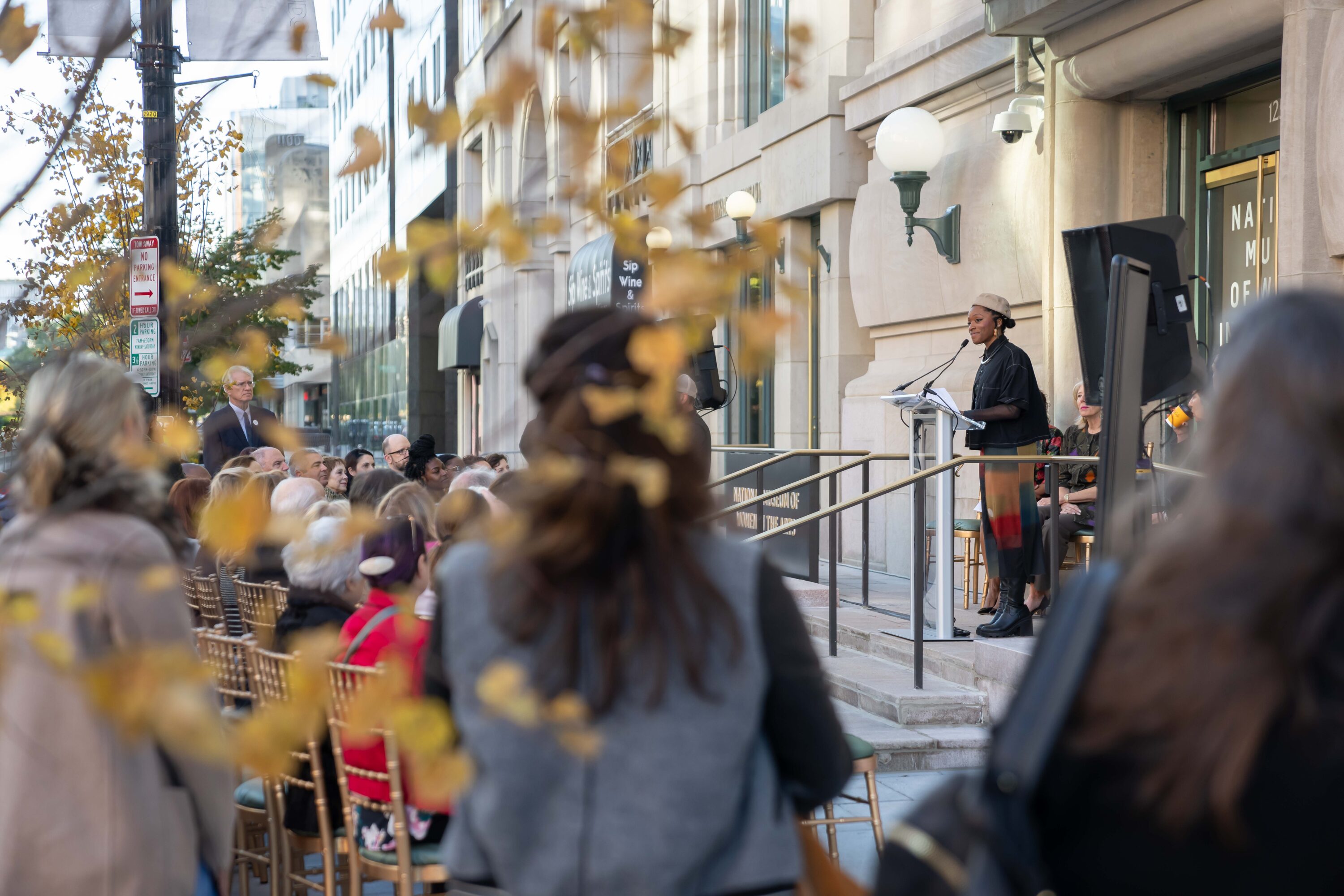 A dark-skinned young woman stands at a podium at the entrance of the National Museum of Women in the Arts, she smiles at a crowd of people who sit in gold-painted chairs on the sidewalk in front of the building.