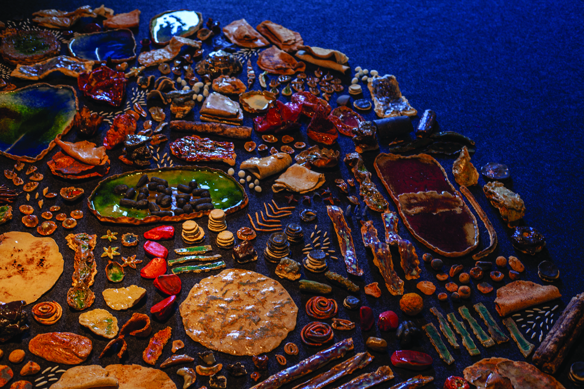 A sculptural arrangement of small, highly-detailed foods made out of ceramic are spaced evenly on the floor in the shape of a circle. The foods are glossy, with lots of textures and warm colors.