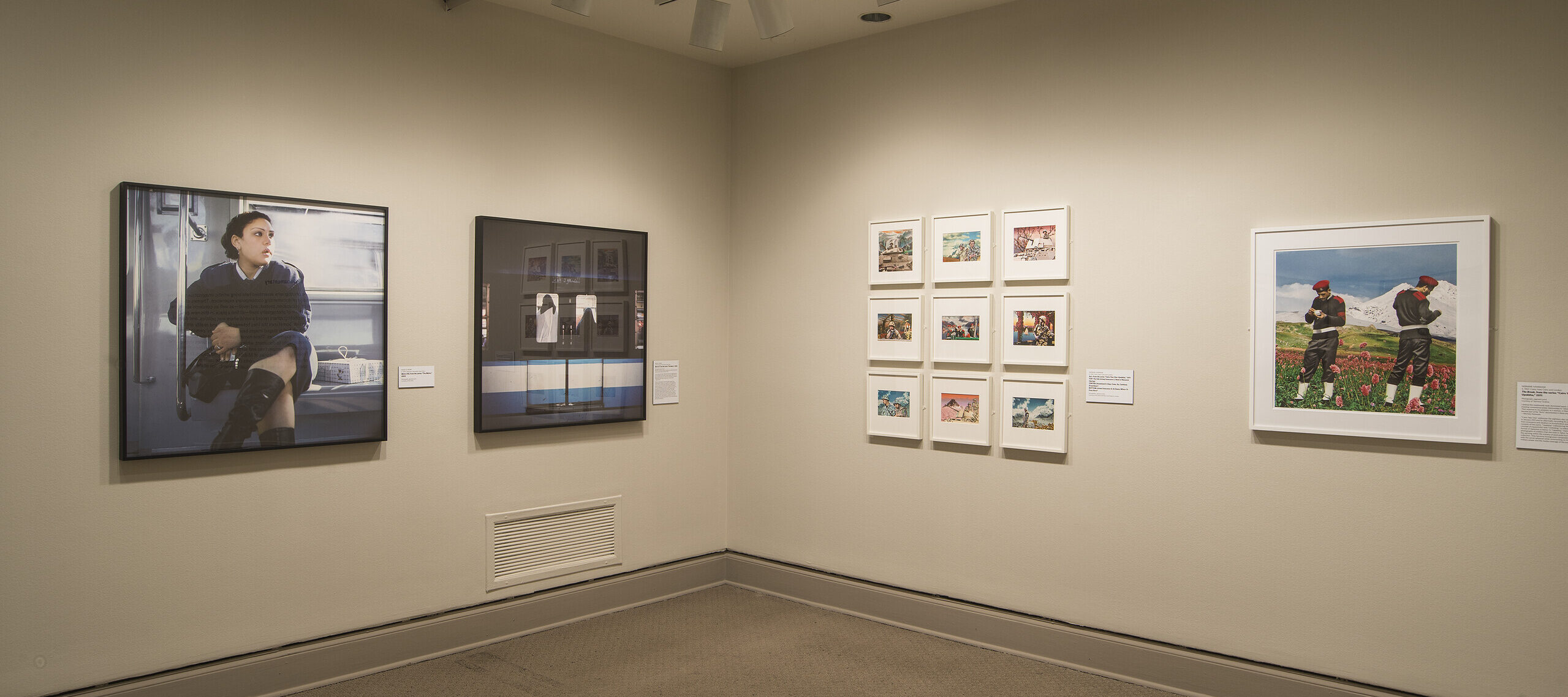 A view of a gallery space. Large prints of photographs are hanging on each wall, depicting a woman in a train and two men in an alpine scene.