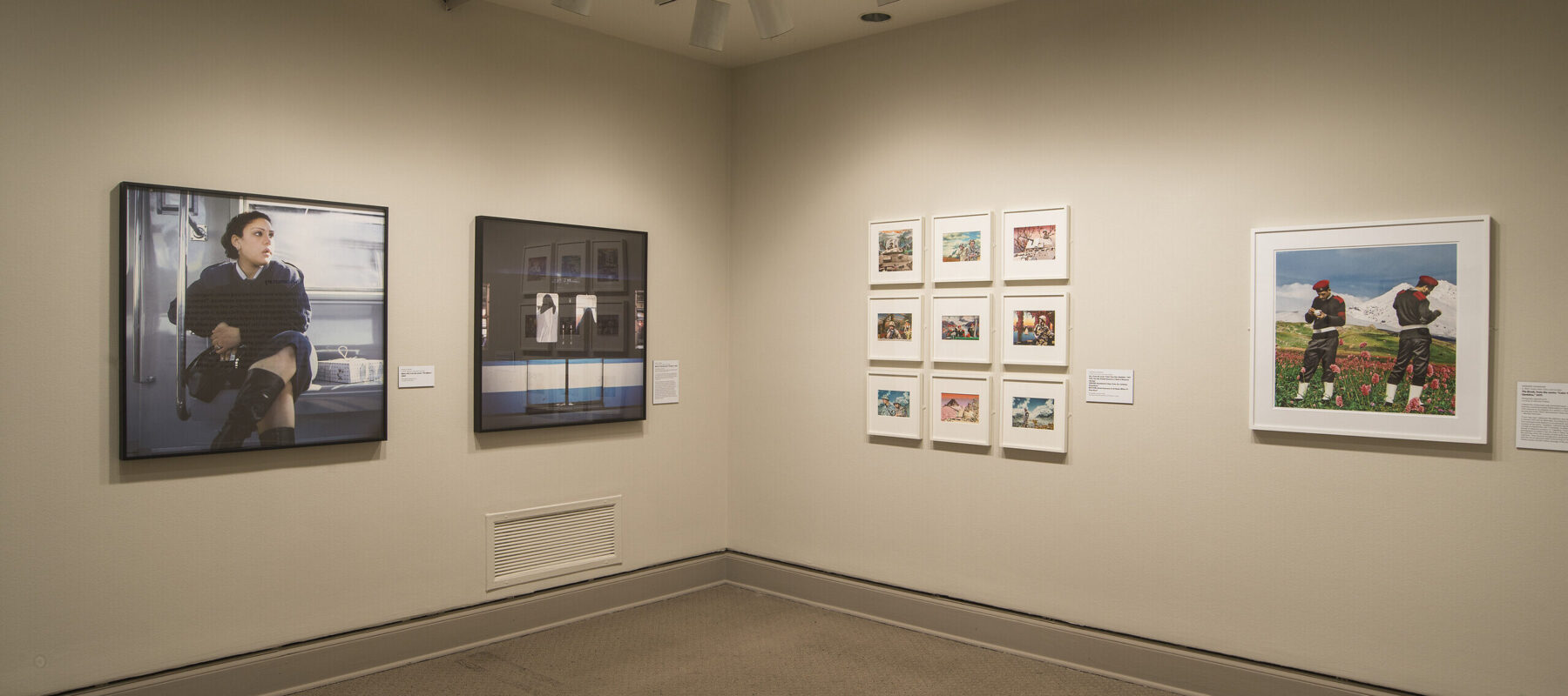 A view of a gallery space. Large prints of photographs are hanging on each wall, depicting a woman in a train and two men in an alpine scene.