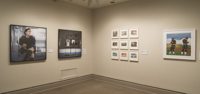 A view of a gallery space. Large prints of photographs are hanging on each wall, depicting a woman in a train and two men in an alpine scene.