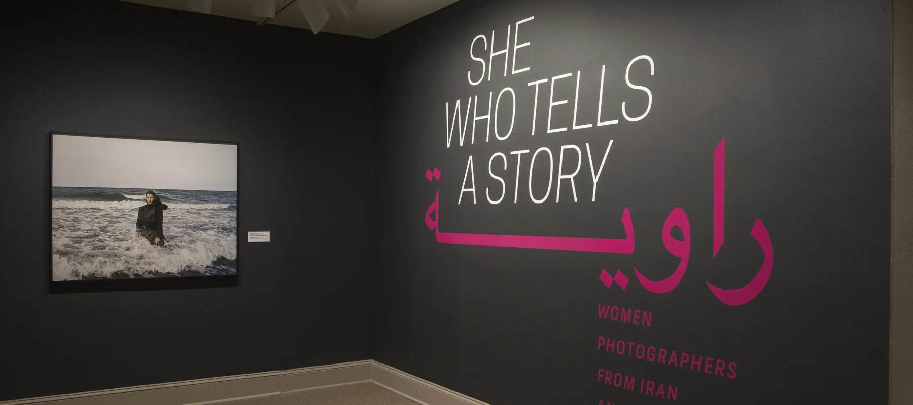 A gallery view of a black wall with a large photograph of a woman. The woman is wearing a long black dress and a head scarf. She is standing in the ocean, surrounded by waves. On the right wall is a text that says "She who tells a story".