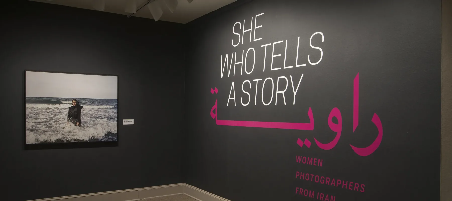 A gallery view of a black wall with a large photograph of a woman. The woman is wearing a long black dress and a head scarf. She is standing in the ocean, surrounded by waves. On the right wall is a text that says "She who tells a story".