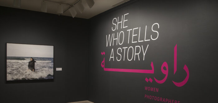 A gallery view of a black wall with a large photograph of a woman. The woman is wearing a long black dress and a head scarf. She is standing in the ocean, surrounded by waves. On the right wall is a text that says "She who tells a story".
