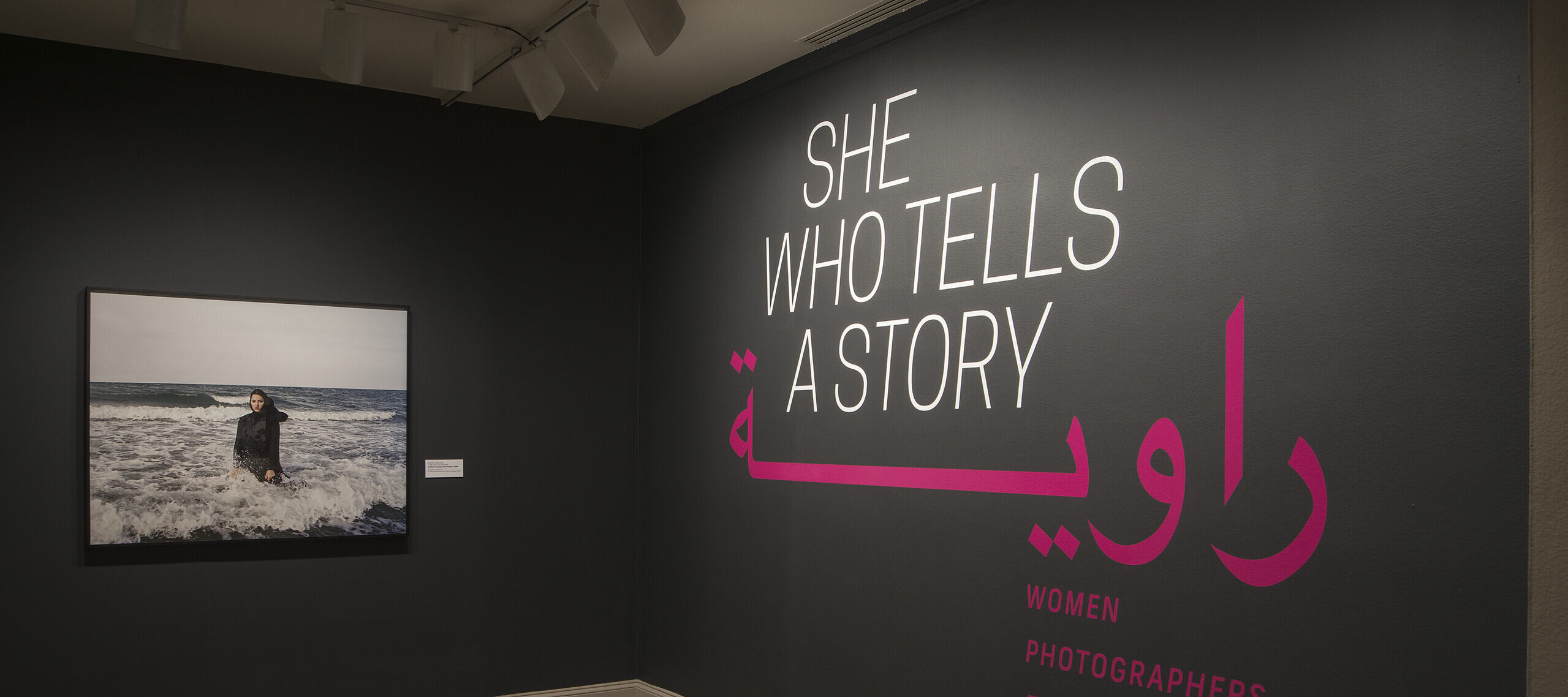 A gallery view of a black wall with a large photograph of a woman. The woman is wearing a long black dress and a head scarf. She is standing in the ocean, surrounded by waves. On the right wall is a text that says 