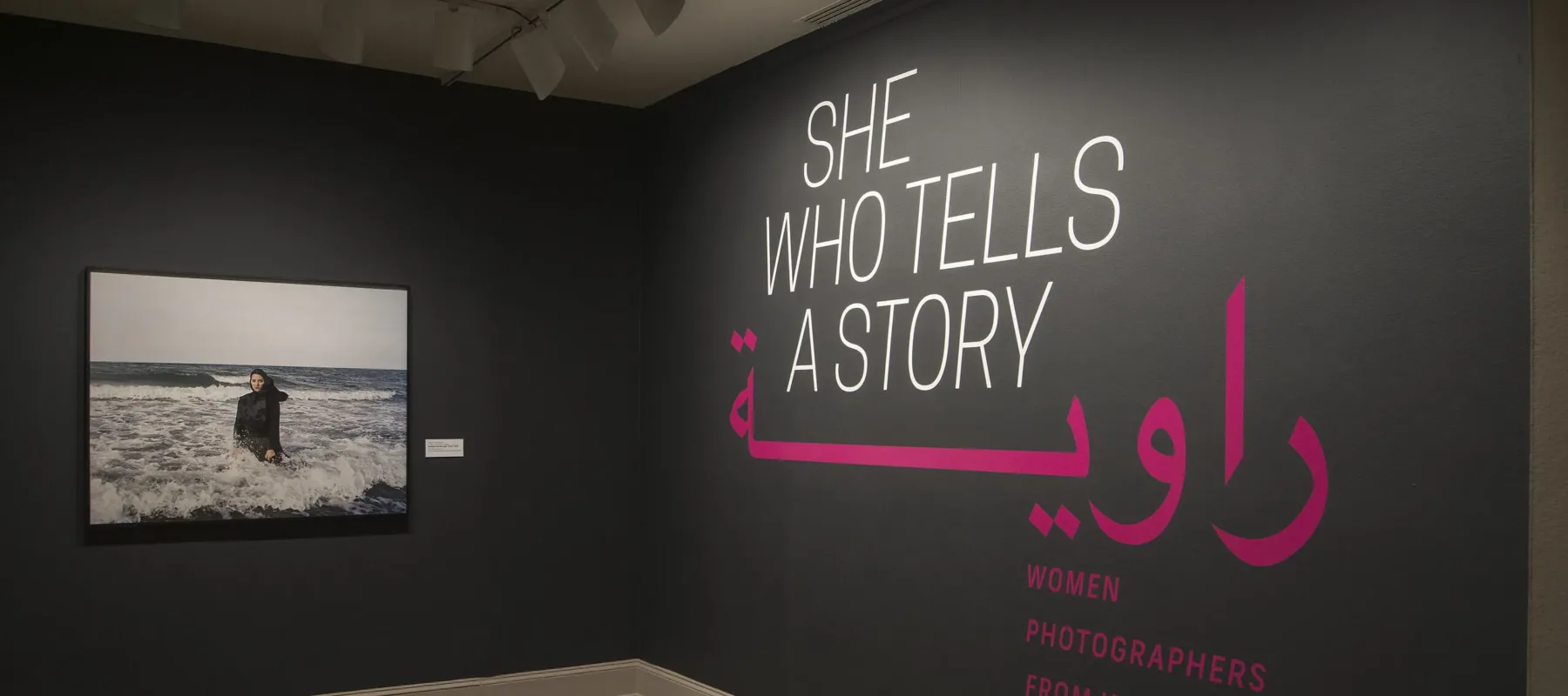 A gallery view of a black wall with a large photograph of a woman. The woman is wearing a long black dress and a head scarf. She is standing in the ocean, surrounded by waves. On the right wall is a text that says "She who tells a story".