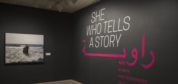 A gallery view of a black wall with a large photograph of a woman. The woman is wearing a long black dress and a head scarf. She is standing in the ocean, surrounded by waves. On the right wall is a text that says "She who tells a story".