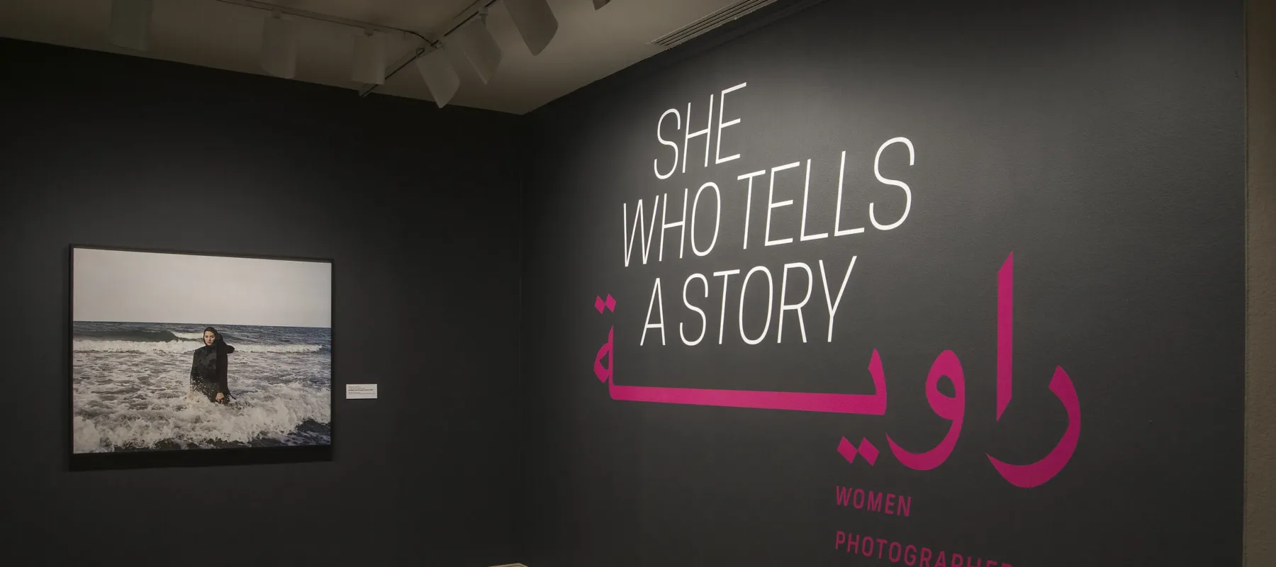 A gallery view of a black wall with a large photograph of a woman. The woman is wearing a long black dress and a head scarf. She is standing in the ocean, surrounded by waves. On the right wall is a text that says "She who tells a story".