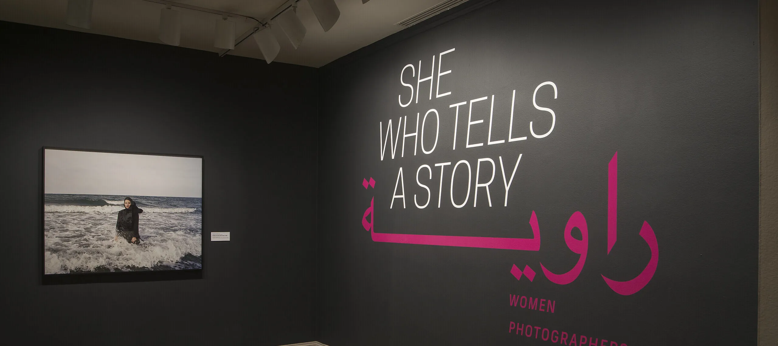 A gallery view of a black wall with a large photograph of a woman. The woman is wearing a long black dress and a head scarf. She is standing in the ocean, surrounded by waves. On the right wall is a text that says "She who tells a story".