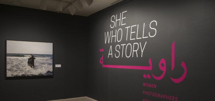 A gallery view of a black wall with a large photograph of a woman. The woman is wearing a long black dress and a head scarf. She is standing in the ocean, surrounded by waves. On the right wall is a text that says "She who tells a story".