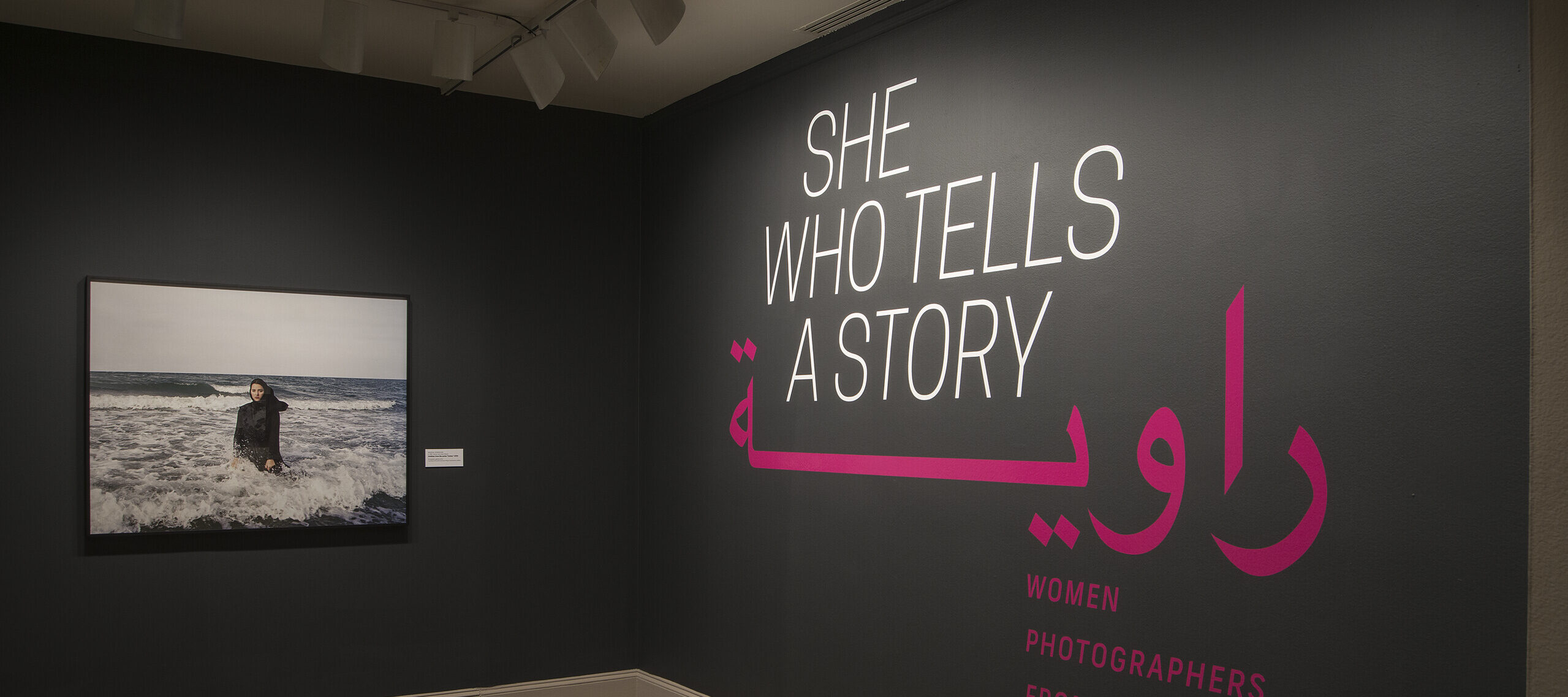 A gallery view of a black wall with a large photograph of a woman. The woman is wearing a long black dress and a head scarf. She is standing in the ocean, surrounded by waves. On the right wall is a text that says 