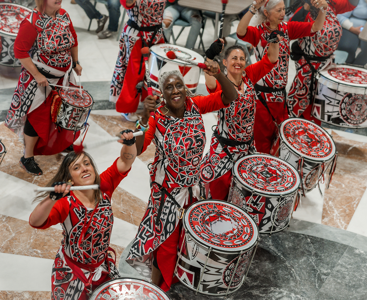 A group of women wearing bright red, black, and white printed dresses play large drums with similar designs on them. They are smiling up at the camera.