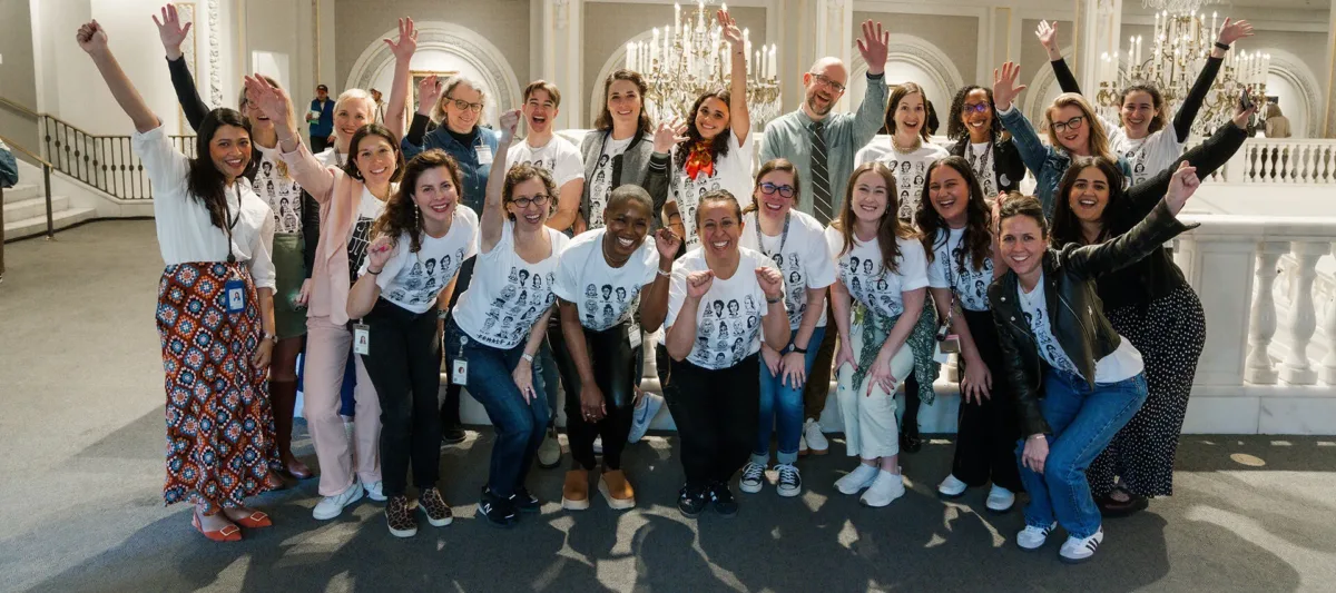 A large group of people wearing the same white-and-black T-shirt pose for a photo in an ornate great hall. They stand in two rows, with the front row people crouching down. Everyone smiles widely and excitedly, some people raise their hands in celebration.