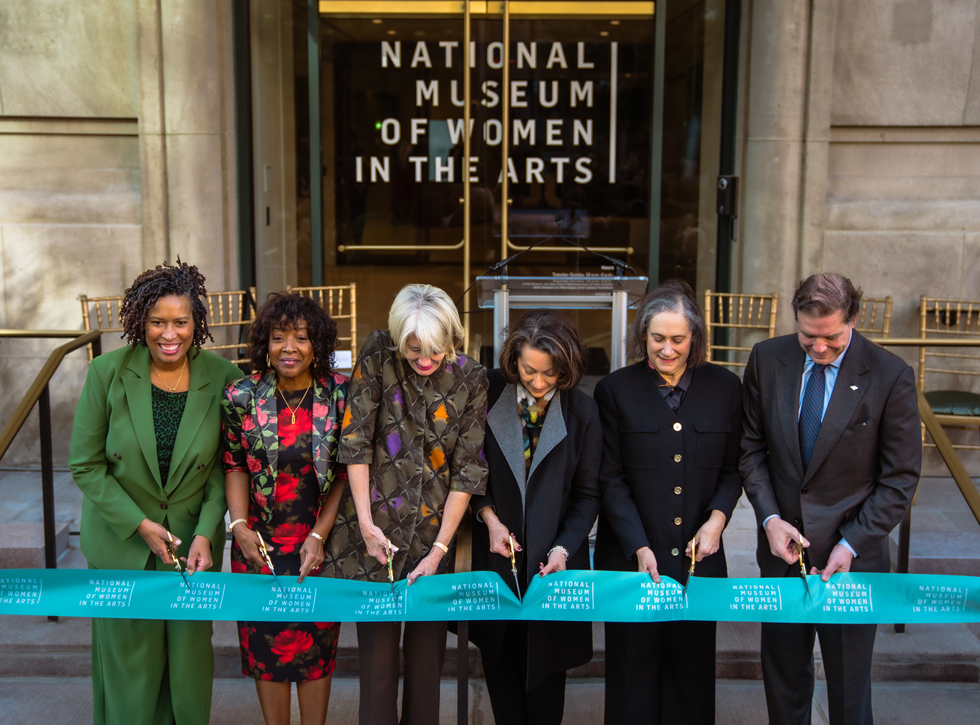 Six people stand in behind a wide green ribbon with scissors, ready to cut it.