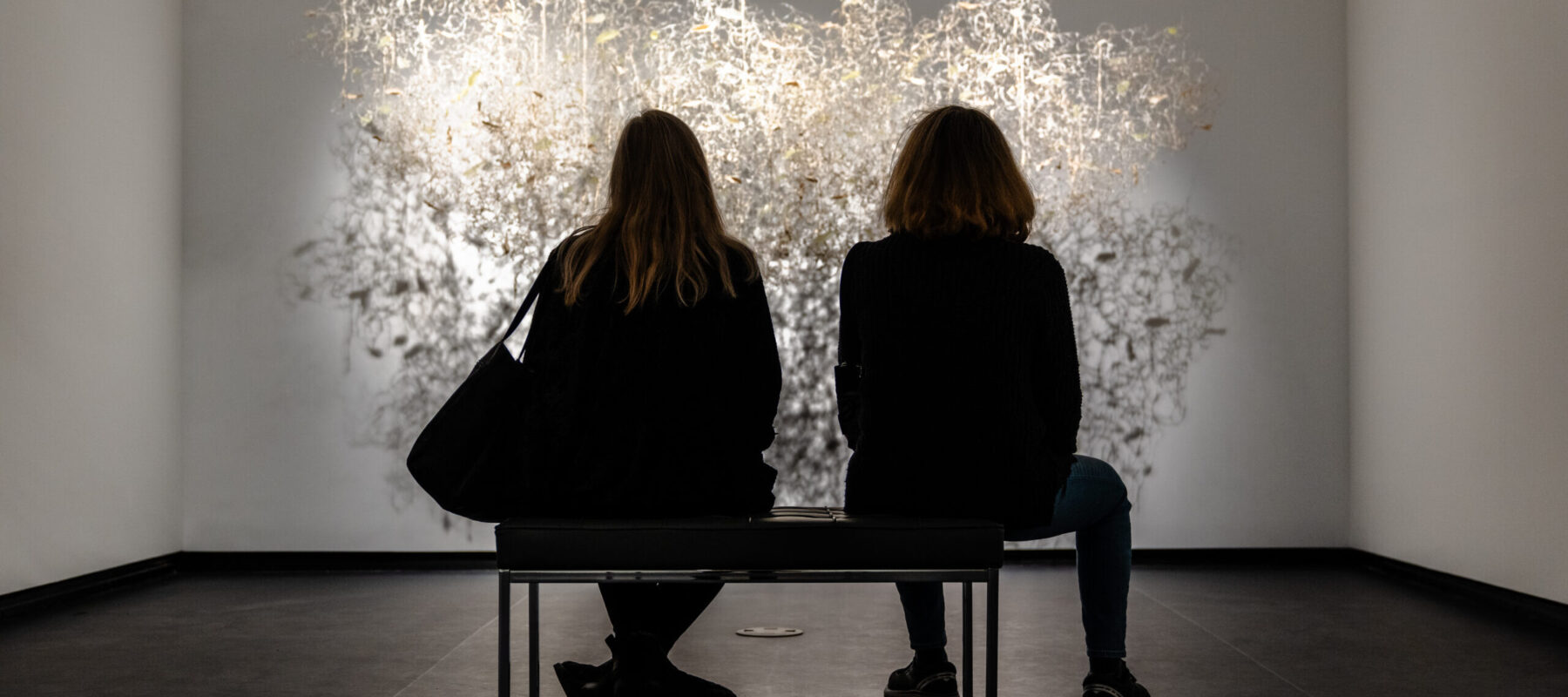 Two museum visitors sit on a bench and observe a brightly lit artwork in a modern gallery.