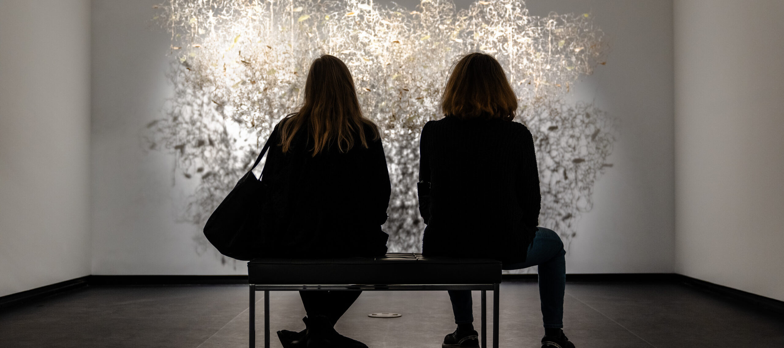 Two museum visitors sit on a bench and observe a brightly lit artwork in a modern gallery.