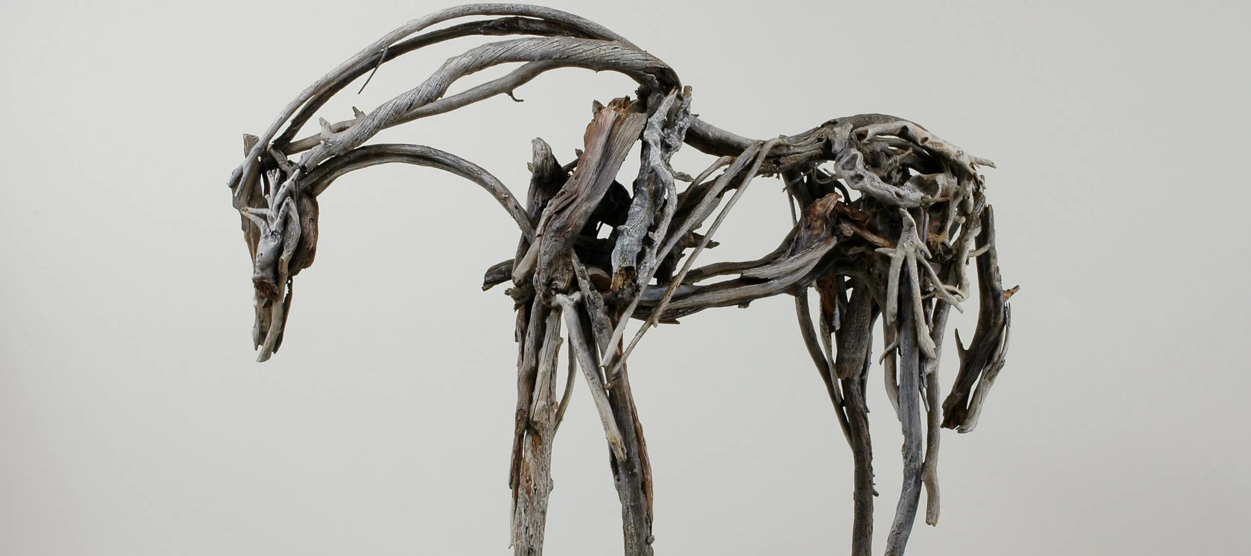 In this life-size sculpture, a horse stands facing left with its neck and head bent down. The horse appears to be made of many sticks of driftwood that are different shades of gray, different textures, and different lengths, all coming together to form a sculpture that evokes a horse’s movement.