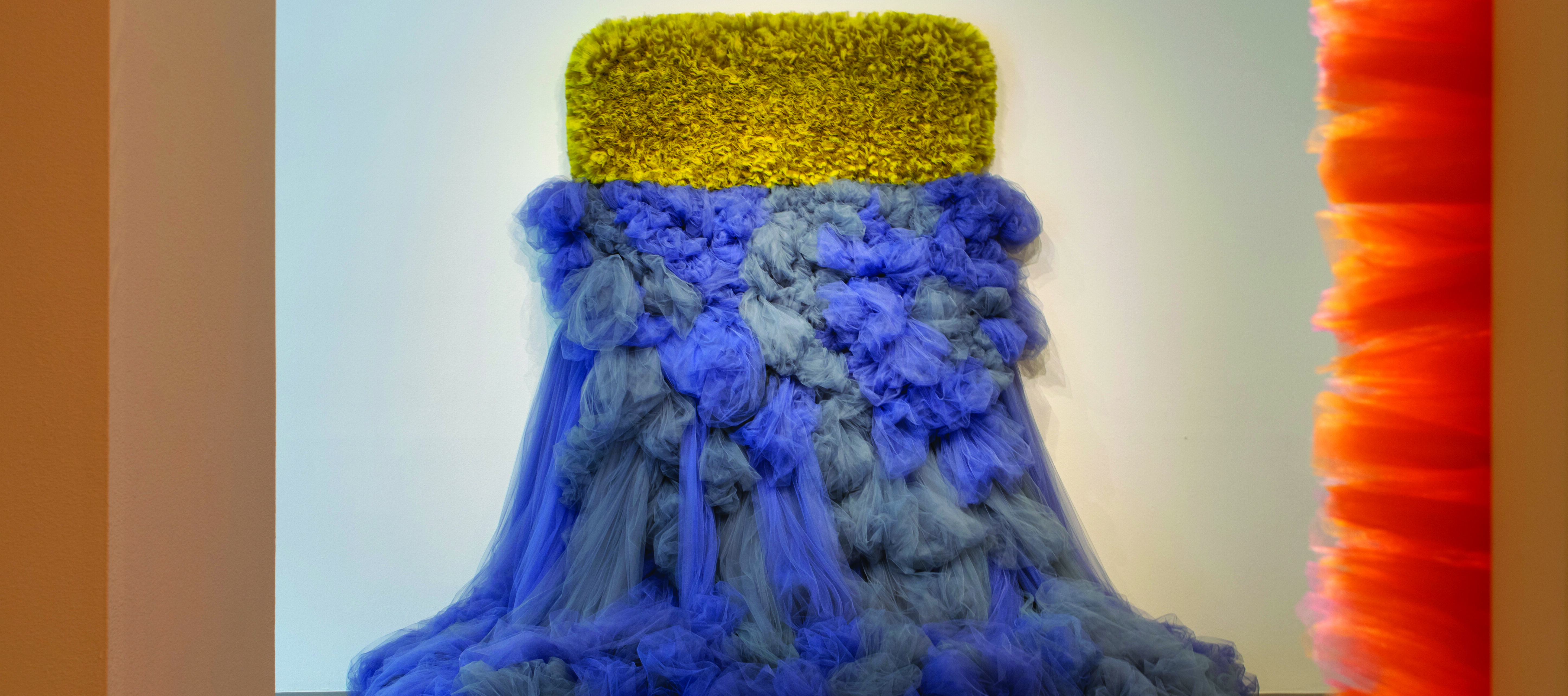 An abstract sculpture made of tulle fabric protrudes from a white wall. The sculpture has two different halves: the tulle on the upper half is cropped close, in a yellow-green horizontal rectangle. The tulle on the lower half is blue-grey, and spills out from the wall onto the floor, like a dress.
