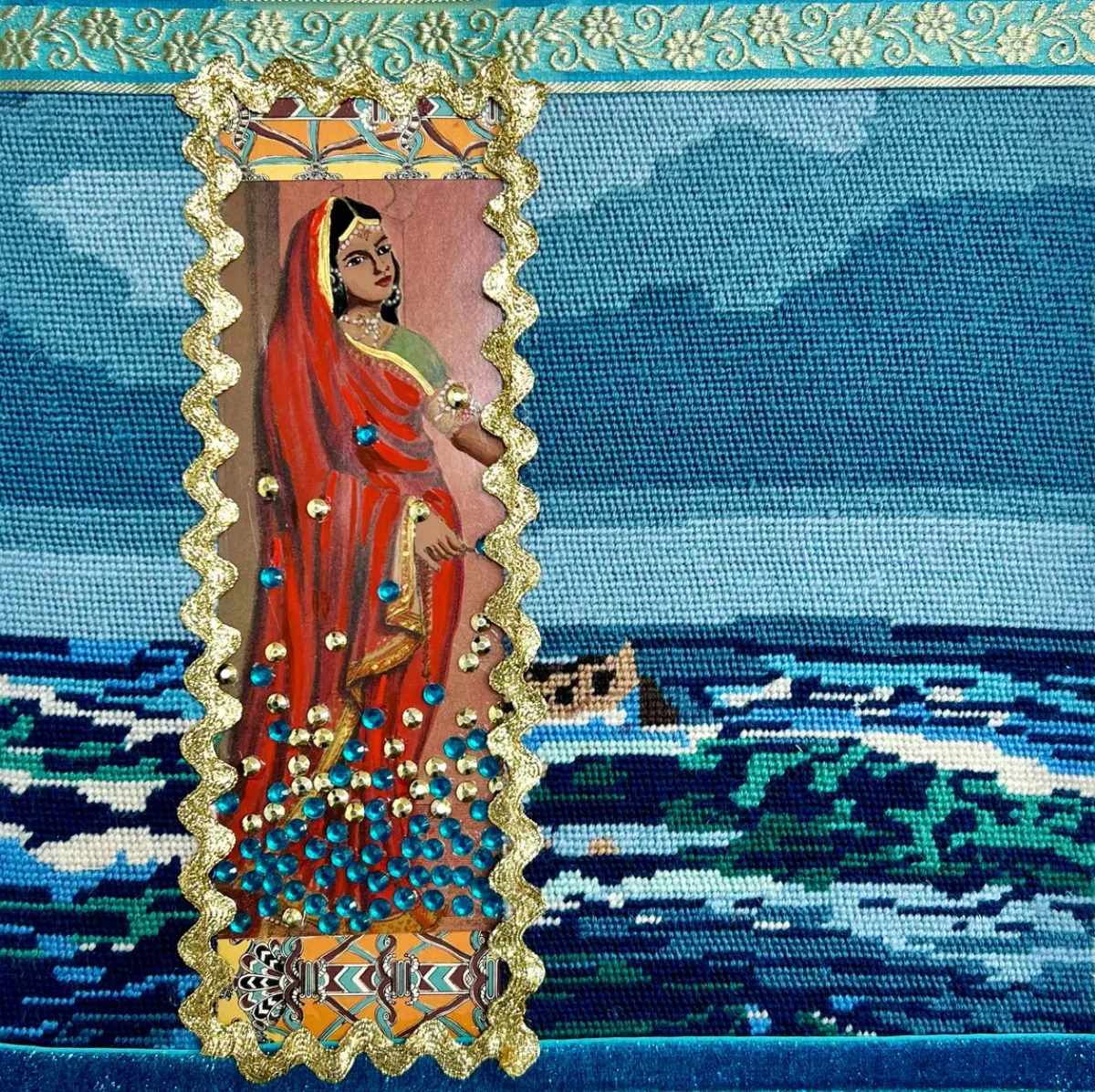 A mixed media work that uses a vintage needlepoint depicting the ocean and sky as its base. Layered over the needlepoint is an image of a standing dark-haired woman wearing a sari. Her lower half is covered in blue and gold rhinestones.