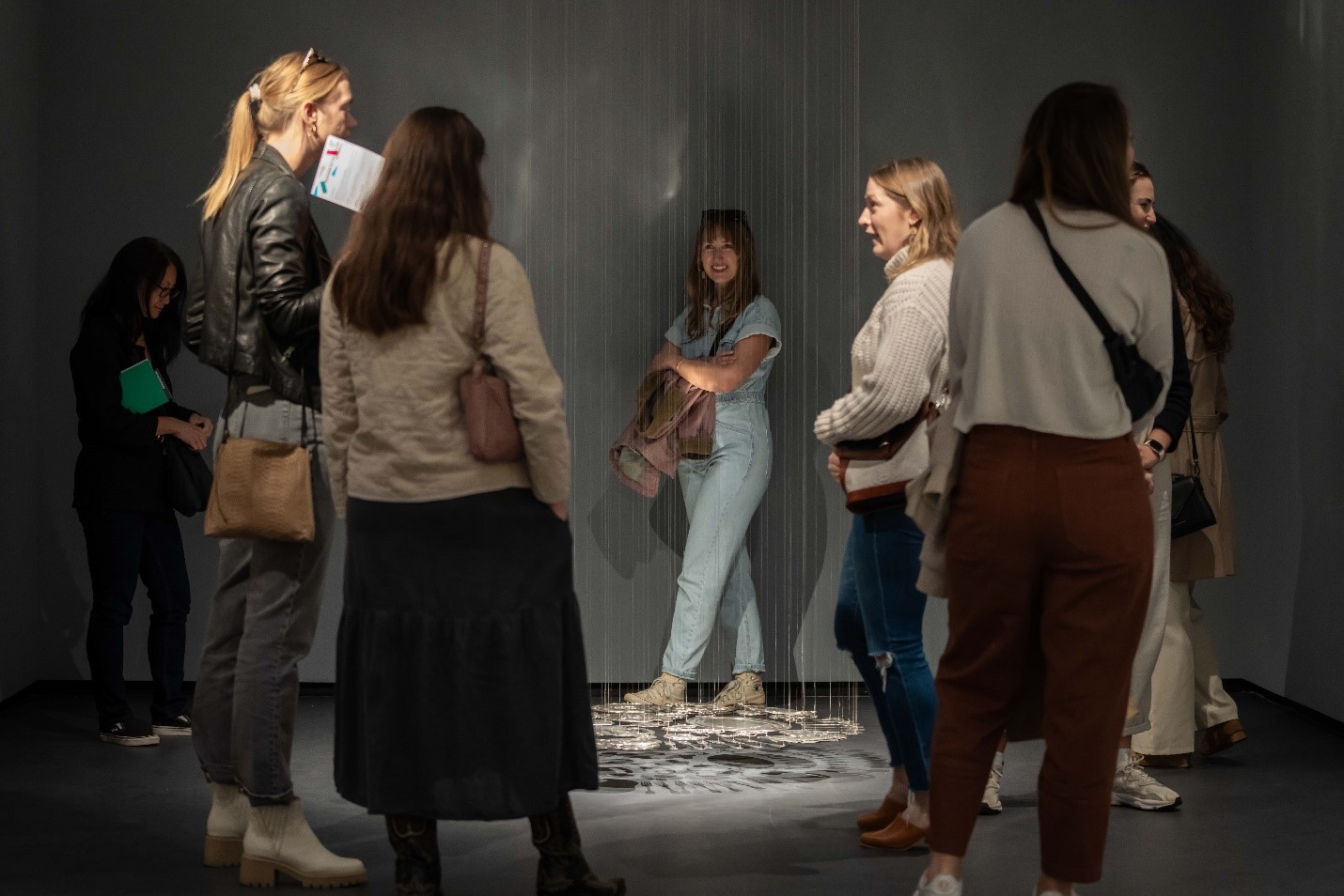 In a dim-lit museum gallery many women walk around a sculpture of flattened silverware that hangs from the ceiling, hovering just above the gallery floor.