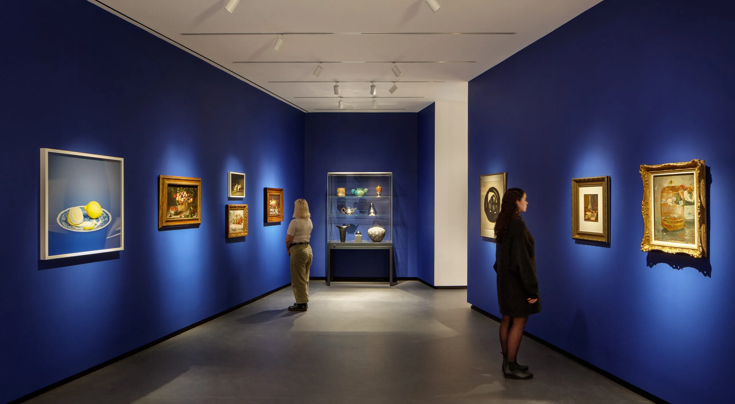 Museum visitors observe artworks hanging on the royal blue walls of a small gallery bay. Paintings of various sizes are on display, and small sculptures are in a glass case.