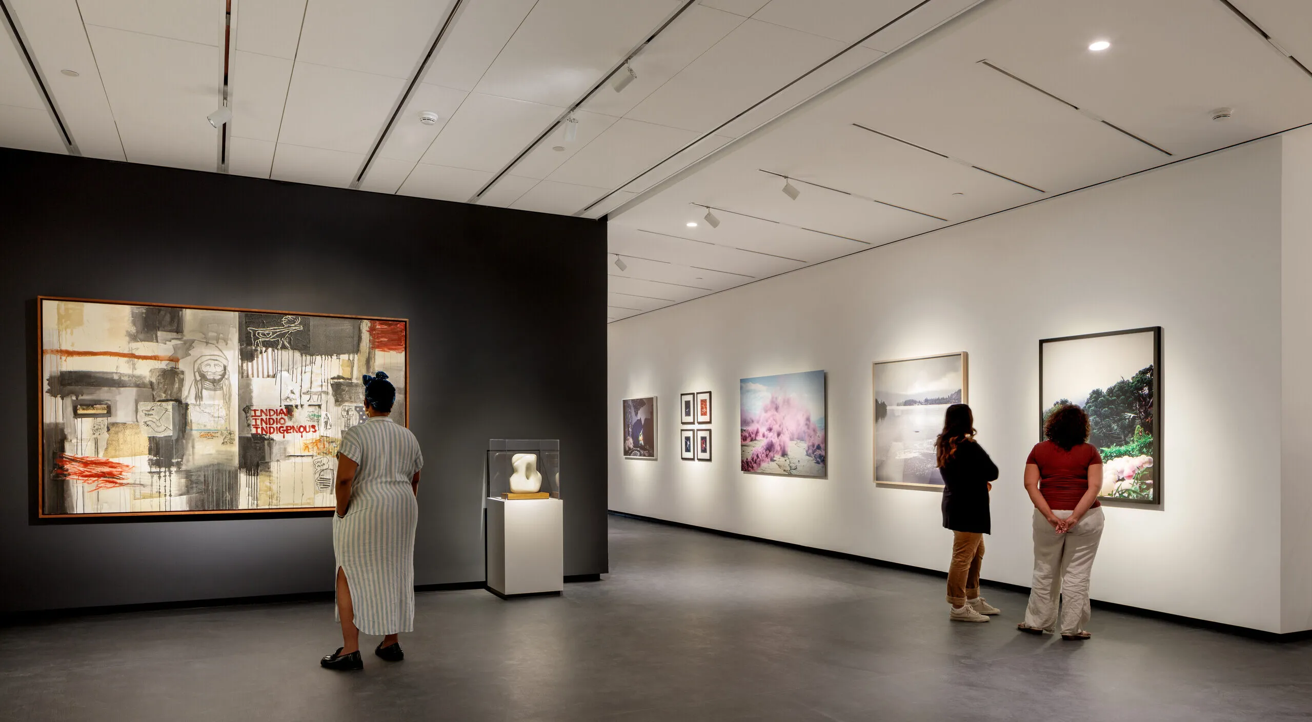 A modern museum gallery is photographed at a wide angle. Visitors observe large artworks on the walls.