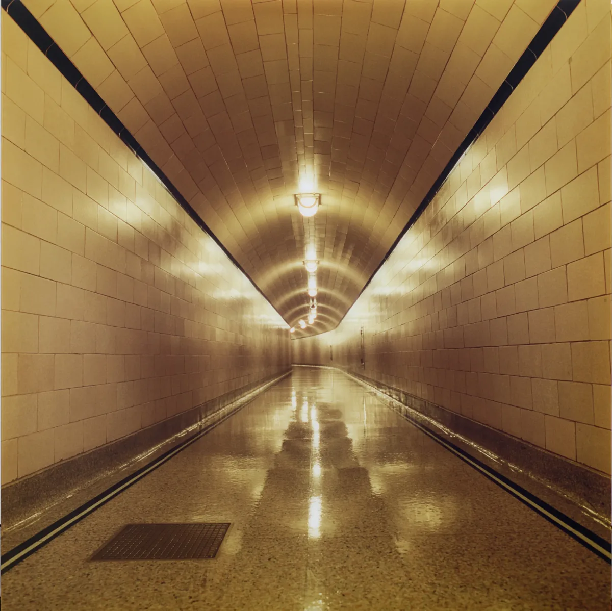 A long, empty corridor with white-tiled walls, floors, and ceiling is lit with a warm glow from the lights that hang in the middle of the ceiling.