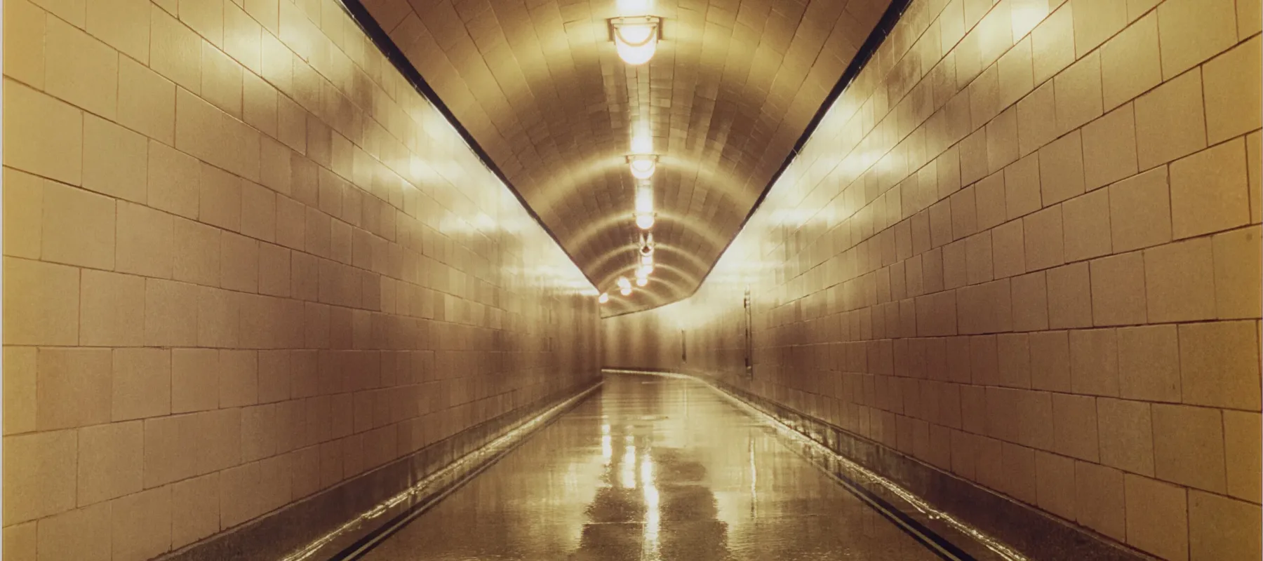 A long, empty corridor with white-tiled walls, floors, and ceiling is lit with a warm glow from the lights that hang in the middle of the ceiling.