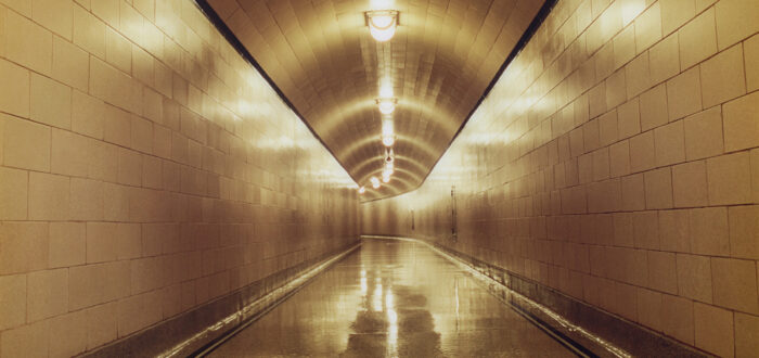 A long, empty corridor with white-tiled walls, floors, and ceiling is lit with a warm glow from the lights that hang in the middle of the ceiling.