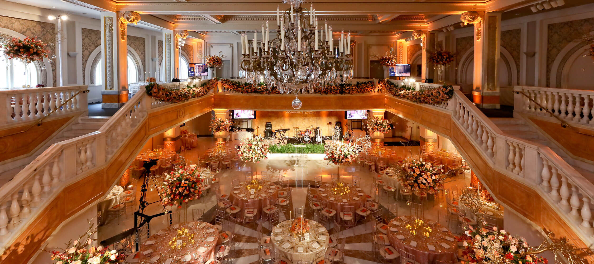 A large two story great hall with a grand staircase and elaborate chandelier, decorated with large floral displays and elegant table settings for a fundraising dinner.
