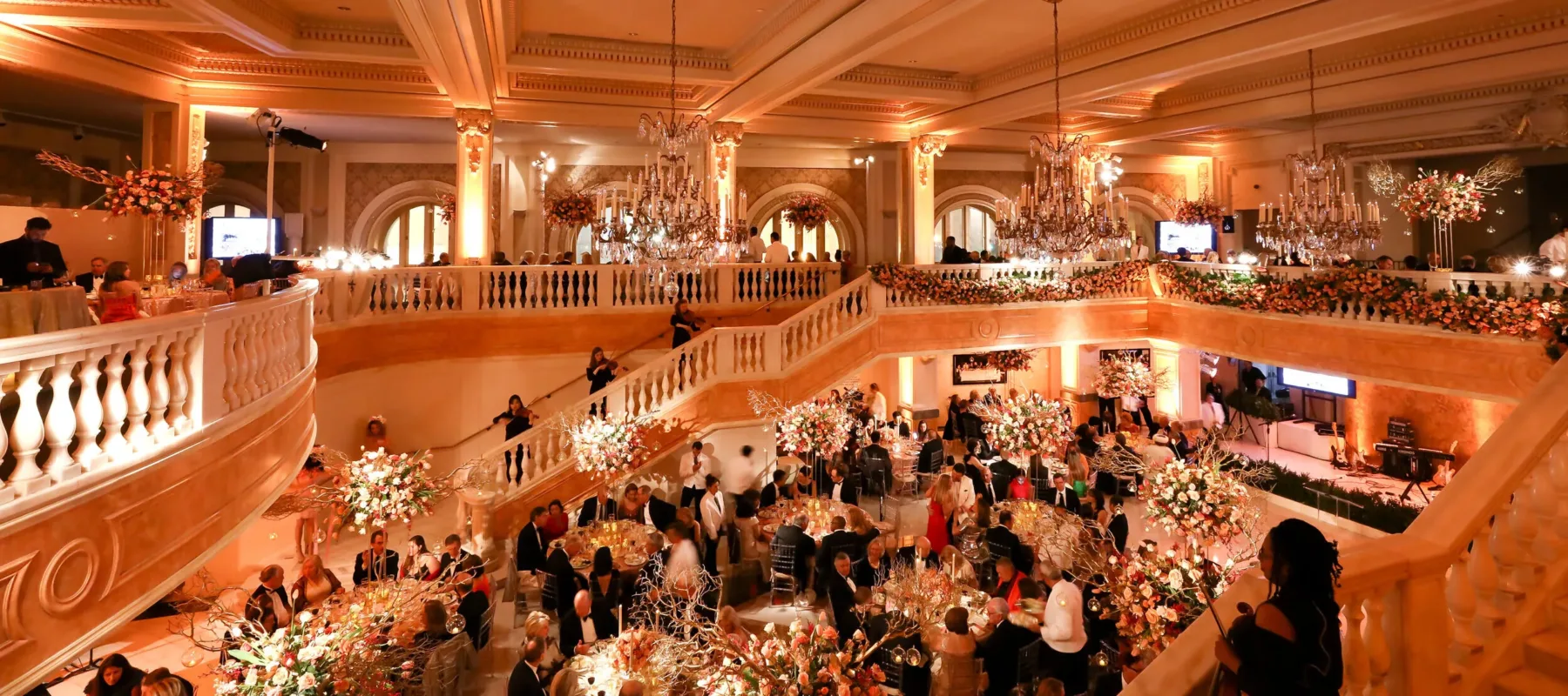 View of the gala from above looking out at the chandeliers, violin players on the marble staircase, and people gathered at round tables surrounded by abundant floral arrangements.