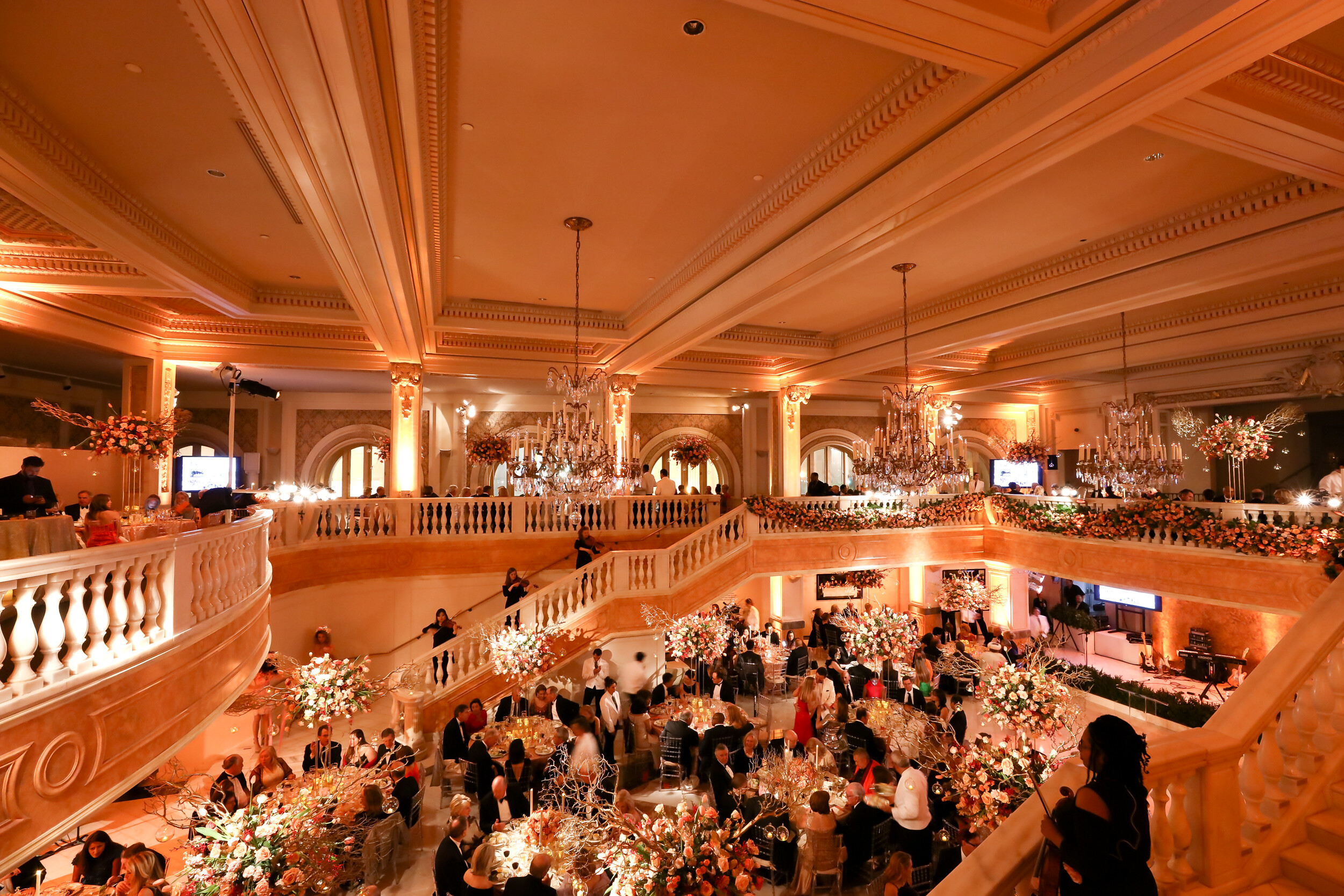 View of the gala from above looking out at the chandeliers, violin players on the marble staircase, and people gathered at round tables surrounded by abundant floral arrangements.