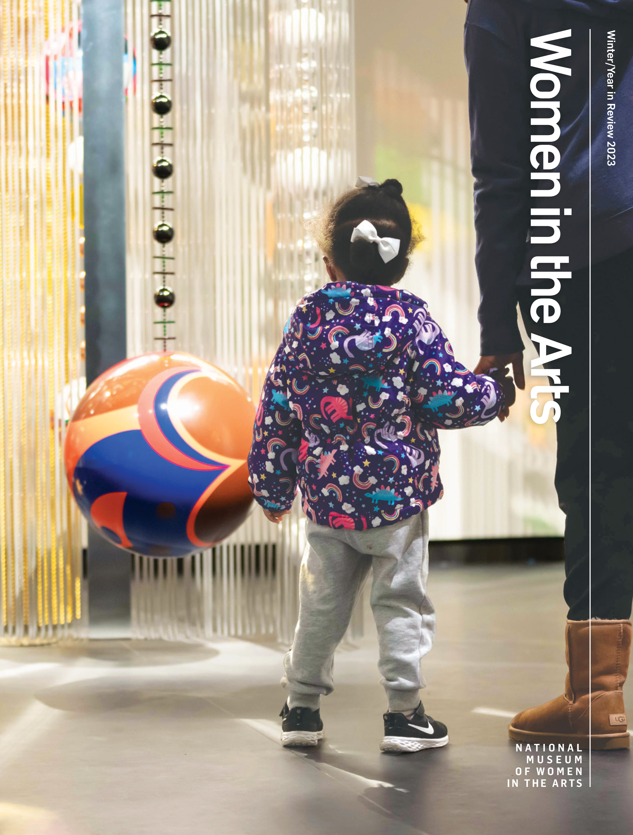 A magazine cover with a photograph of a medium-skinned child wearing a whimsical jacket and white bows in their hair, looking at a 3 dimensional sculpture with a large colorful ball suspended above the floor and long strands of beads hanging around it.
