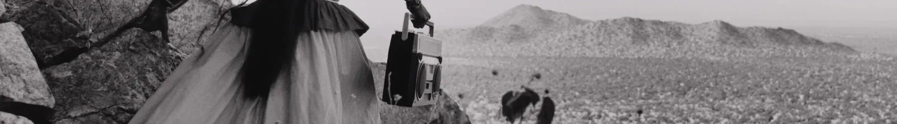 A black-and-white photograph shows the back of a woman as she crests a rocky path above a vast desert landscape beneath an expansive sky. Her traditional, ethnic full skirt, long-sleeved blouse, and long, straight, dark hair contrasts with the modern portable stereo she carries.