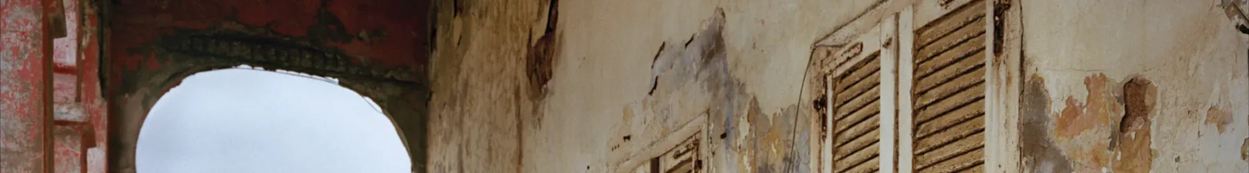 A light-skinned young woman with long, dark brown hair in a black, long lace sleeved dress stands confidently in a crumbling loggia. She gazes at the viewer with a serious, captivating look.