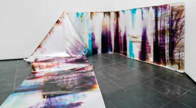 In an art gallery with white walls, a long, snake-like piece of metallic paper printed with splotches of black, blue, purple, and orange in an abstract pattern is partly affixed to the wall. At one point the paper falls from the wall and cascades onto the floor and further into the room.