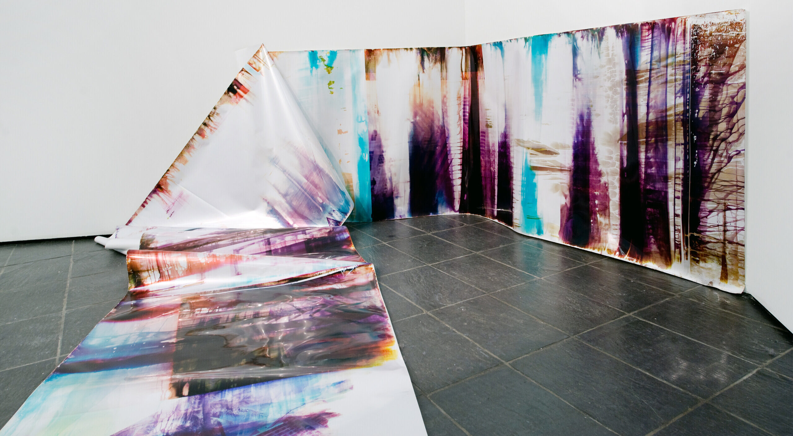 In an art gallery with white walls, a long, snake-like piece of metallic paper printed with splotches of black, blue, purple, and orange in an abstract pattern is partly affixed to the wall. At one point the paper falls from the wall and cascades onto the floor and further into the room.