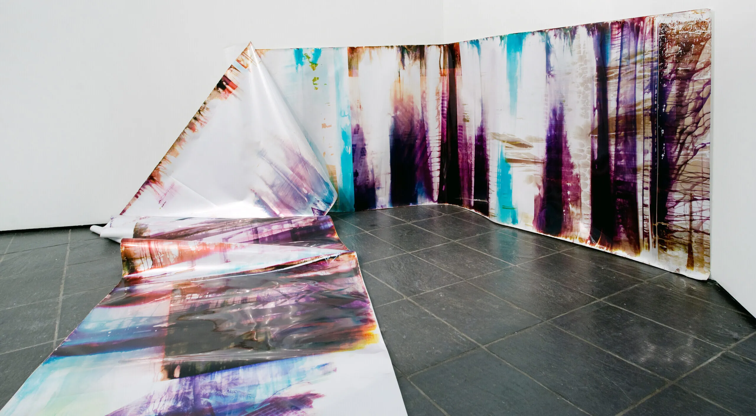 In an art gallery with white walls, a long, snake-like piece of metallic paper printed with splotches of black, blue, purple, and orange in an abstract pattern is partly affixed to the wall. At one point the paper falls from the wall and cascades onto the floor and further into the room.